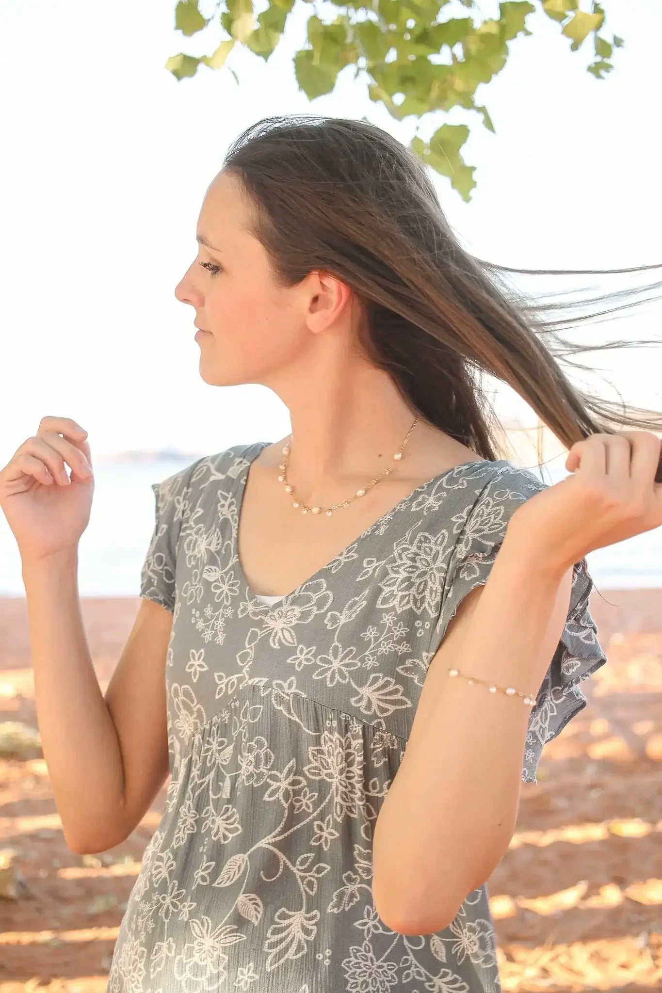 A woman in a floral-patterned gray dress playfully gestures with her hands, while her hair flows gently in a breezy outdoor setting near the water, surrounded by leafy branches and sandy ground.