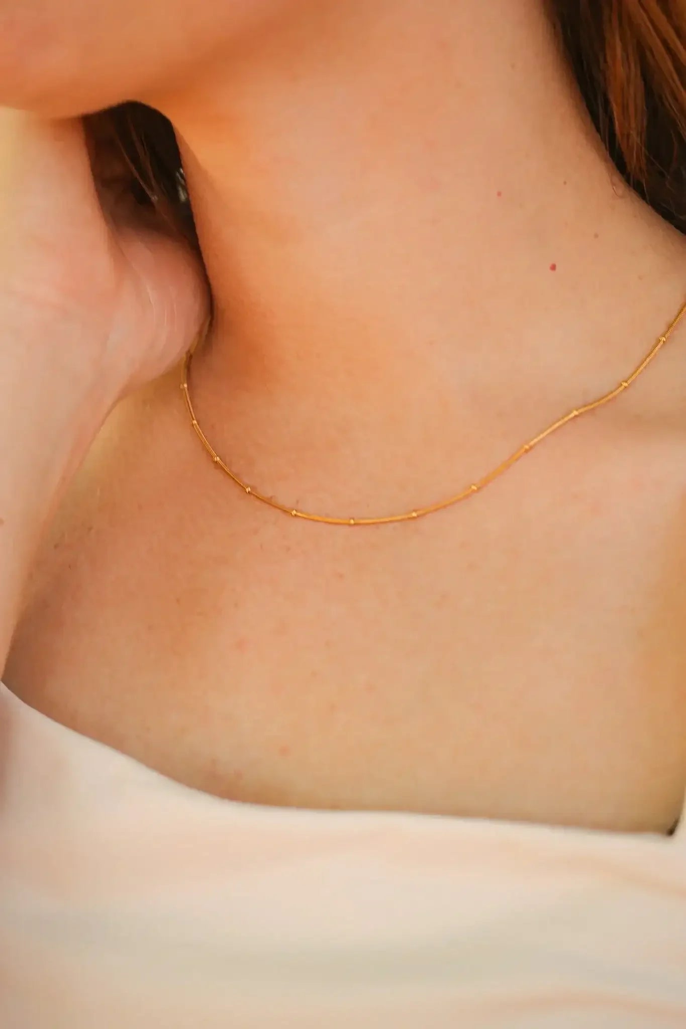 A delicate gold necklace rests on a person's neck, while a hand gently touches the area. The background suggests an outdoor setting with soft lighting.