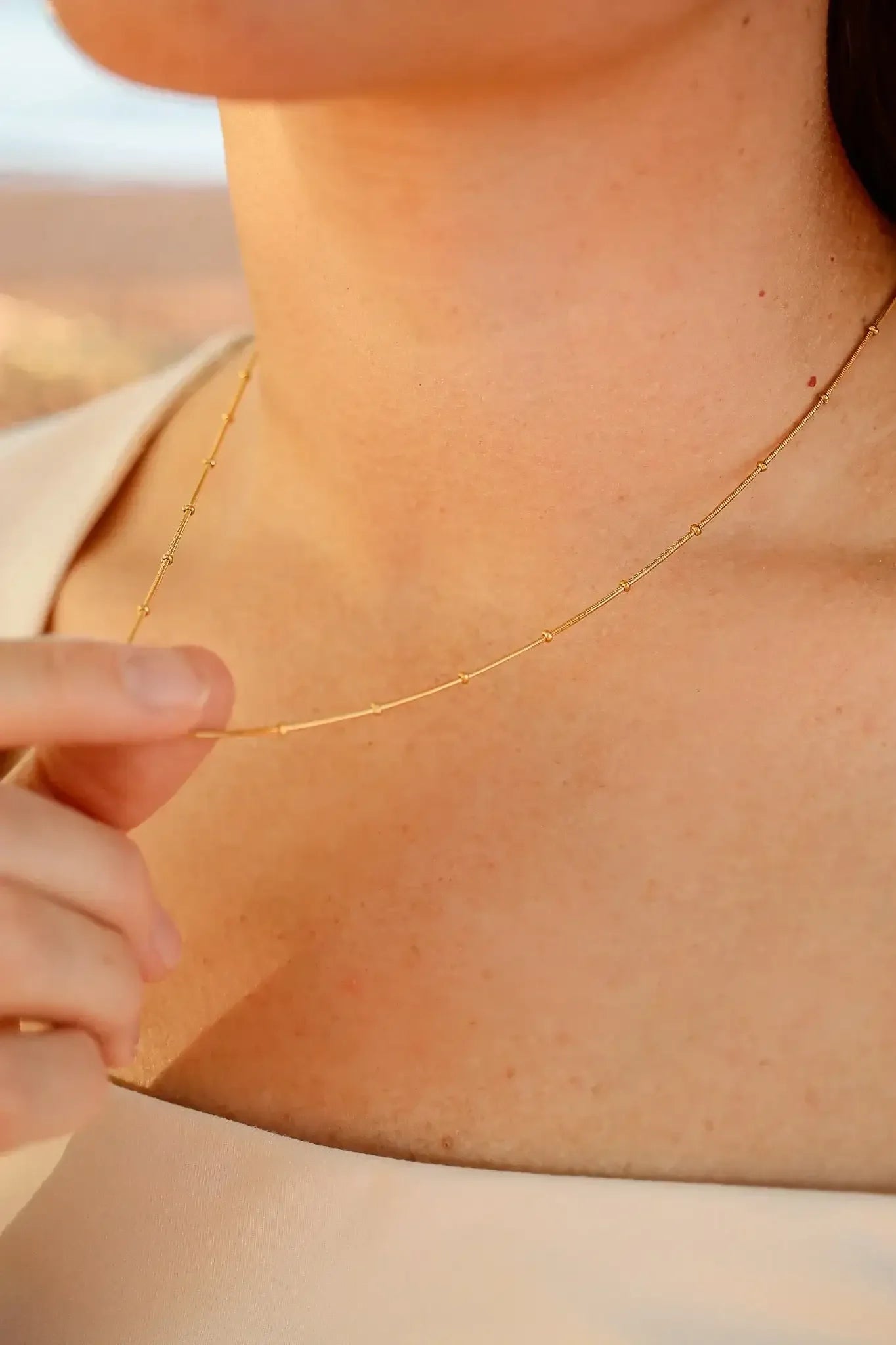 A hand gently holds a delicate gold necklace resting on a woman's neck, complementing her light-colored garment against a soft, blurred outdoor background.