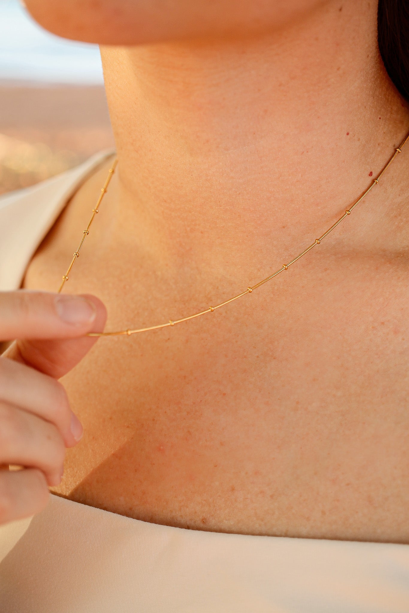 A hand gently holds a delicate gold necklace resting on a woman's neck, complementing her light-colored garment against a soft, blurred outdoor background.