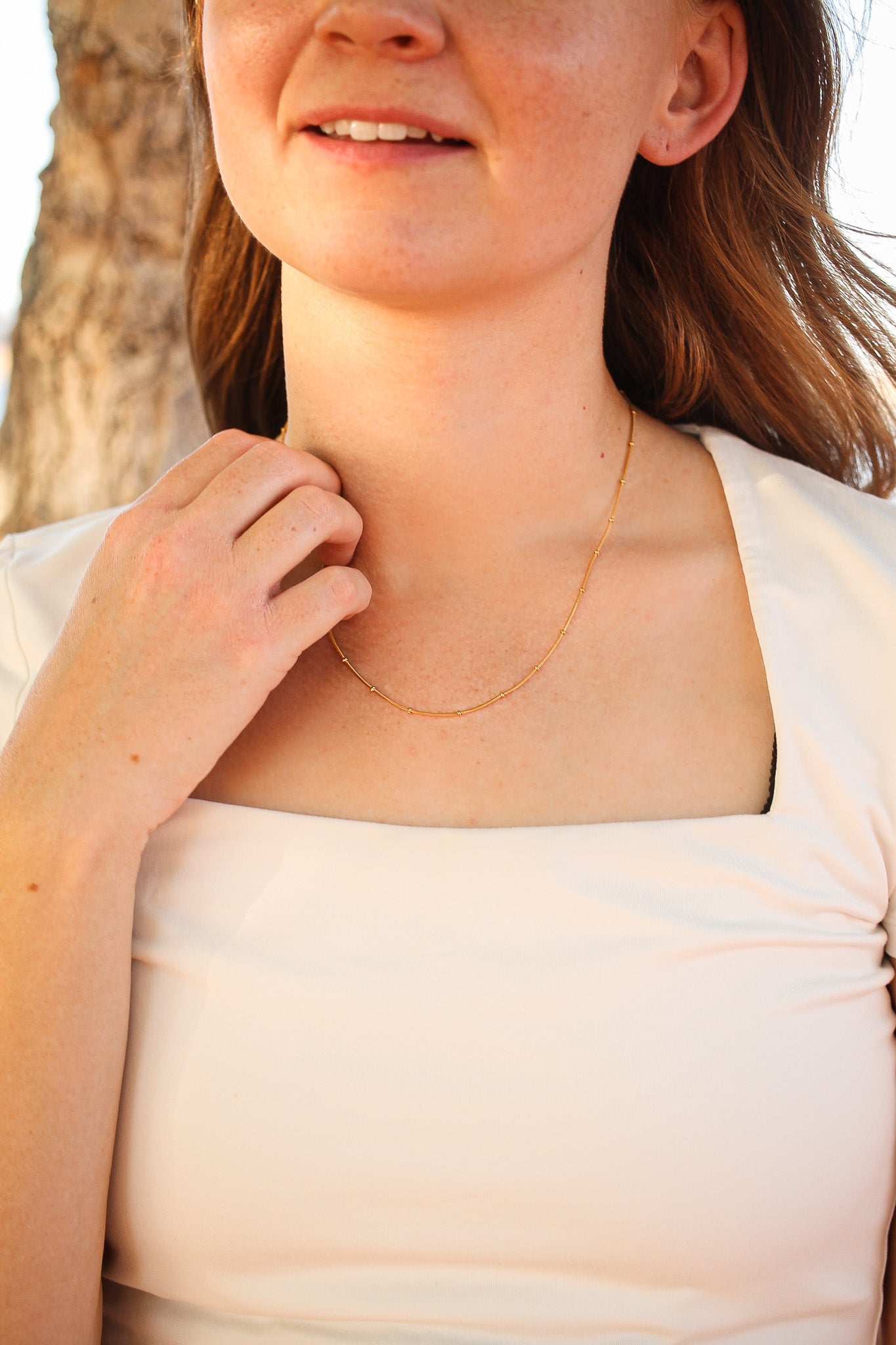 A woman gently touches her necklace, wearing a light-colored top with a square neckline. Natural light highlights her features, indicating an outdoor setting with a soft, blurred background.