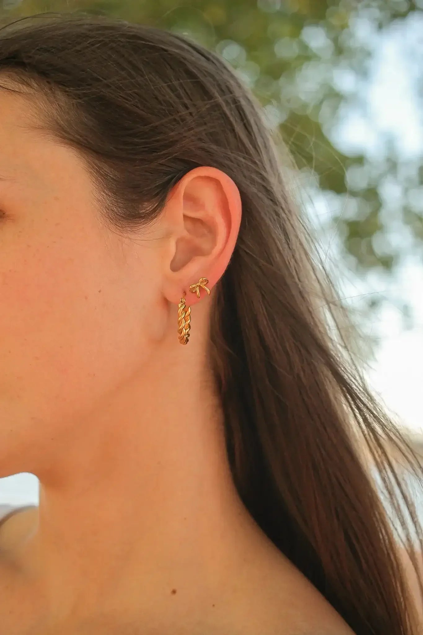 A woman's ear is adorned with two gold earrings: a thin twisted hoop and a small decorative piece shaped like a bow, with soft natural light in the background.