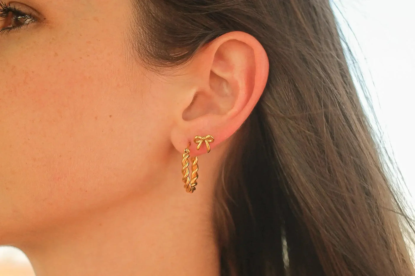 A close-up of a woman's ear displays two gold earrings: a twisted hoop and a small bow-shaped stud. Sunlight highlights her smooth skin and flowing hair in a bright background.
