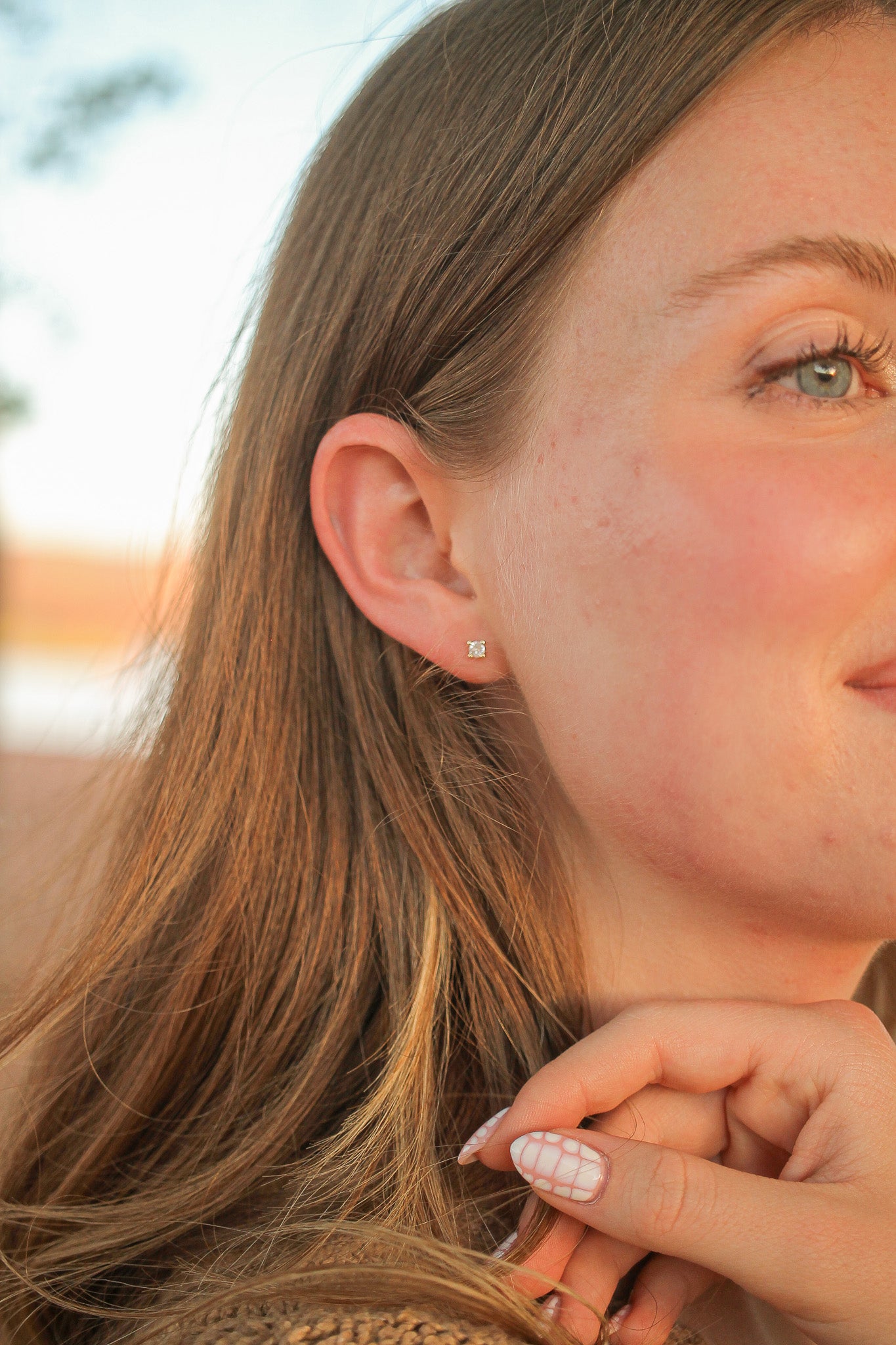 A young woman with light brown hair smiles, touching her ear with one hand. She wears a small stud earring. The background shows a blurred outdoor setting, suggesting a warm atmosphere.