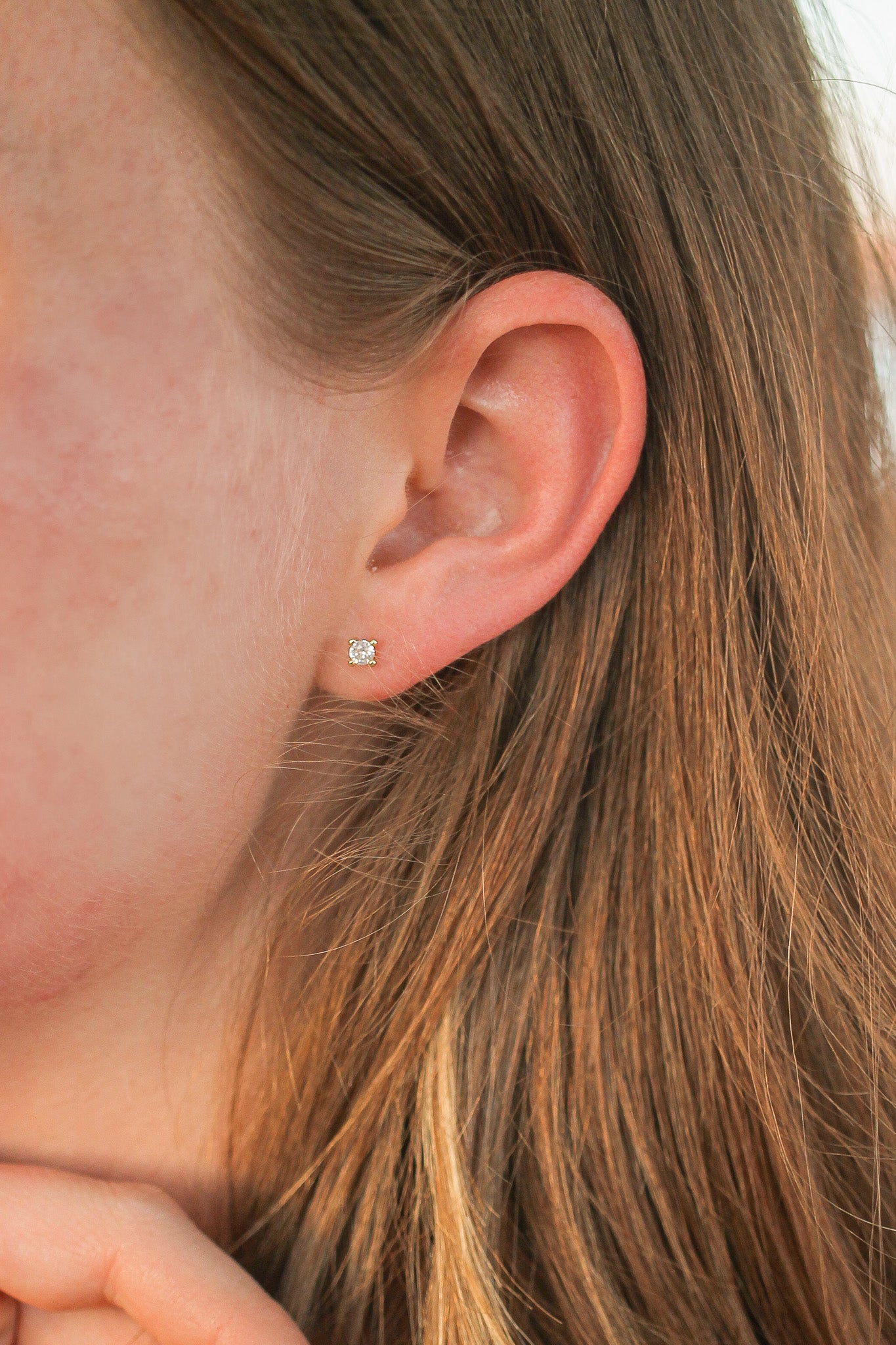 A close-up of an ear featuring a small, sparkling stud earring. The background shows long, wavy brown hair, suggesting a casual outdoor setting with natural lighting.