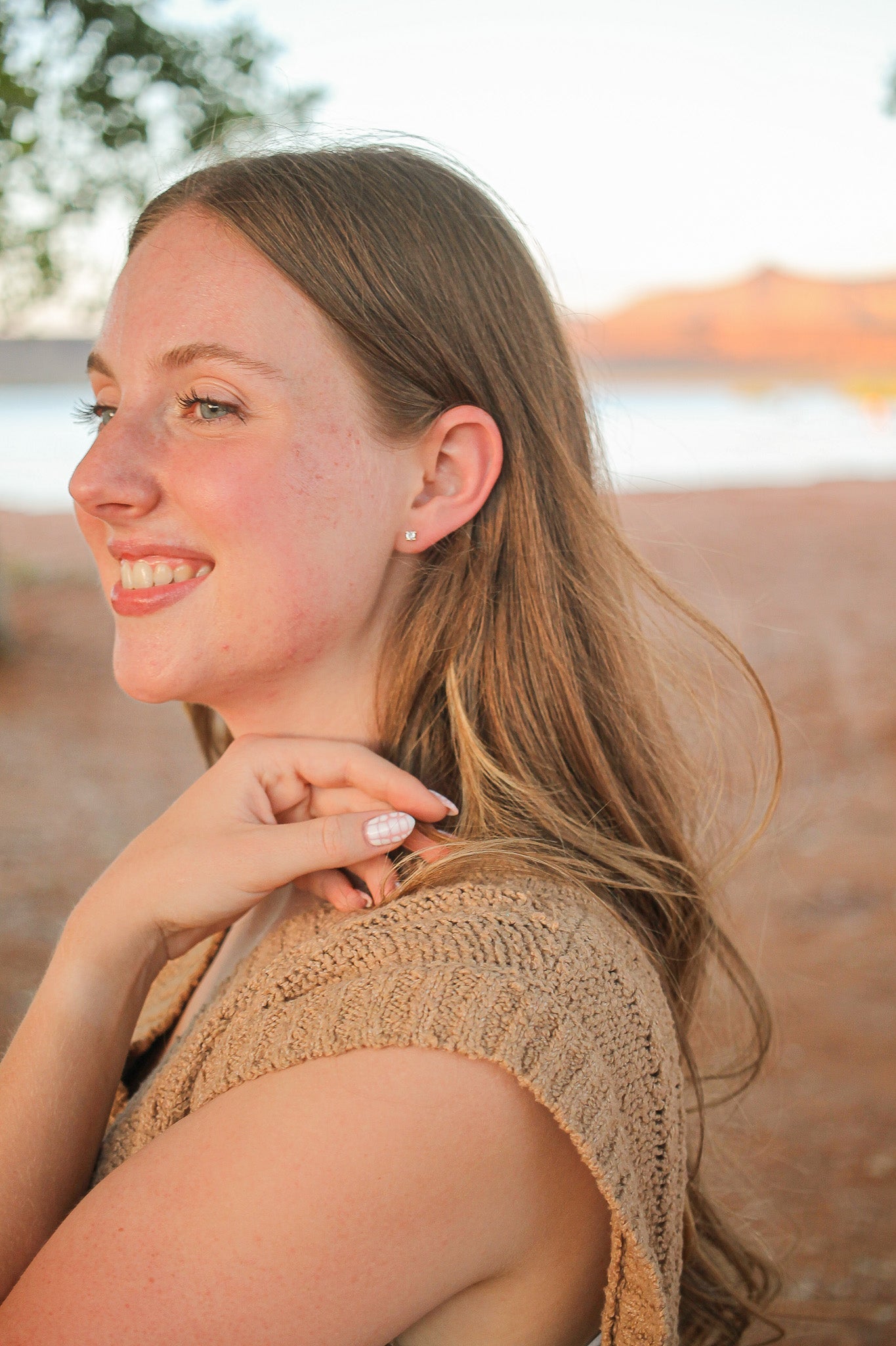 A young woman smiles while touching her hair, wearing a sleeveless, textured beige top. She stands in a natural setting, with a blurred water body and hills in the background.