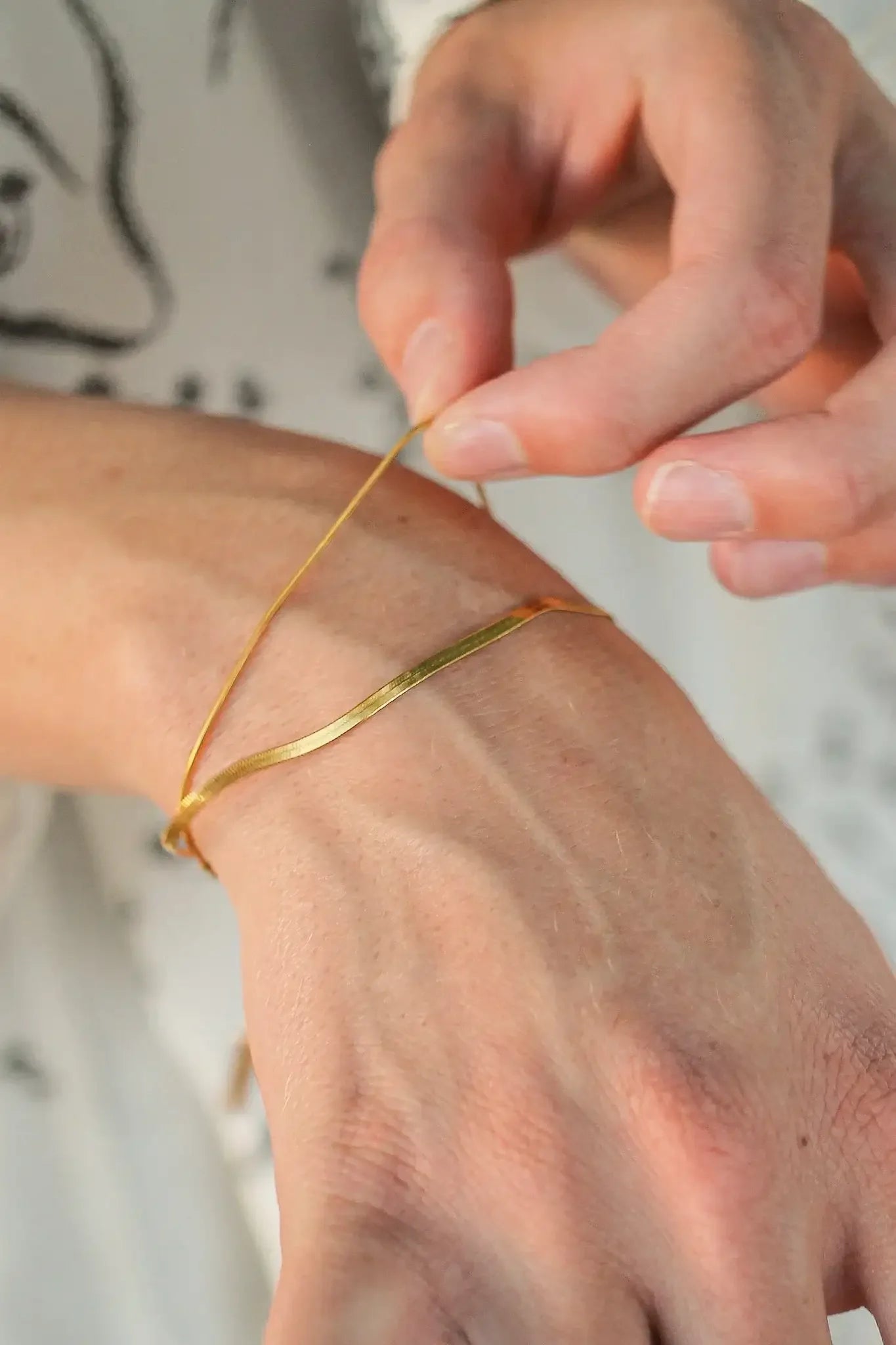 A hand is adjusting a thin gold bracelet around a wrist. The background features light-colored fabric with abstract patterns, creating a soft and elegant atmosphere.