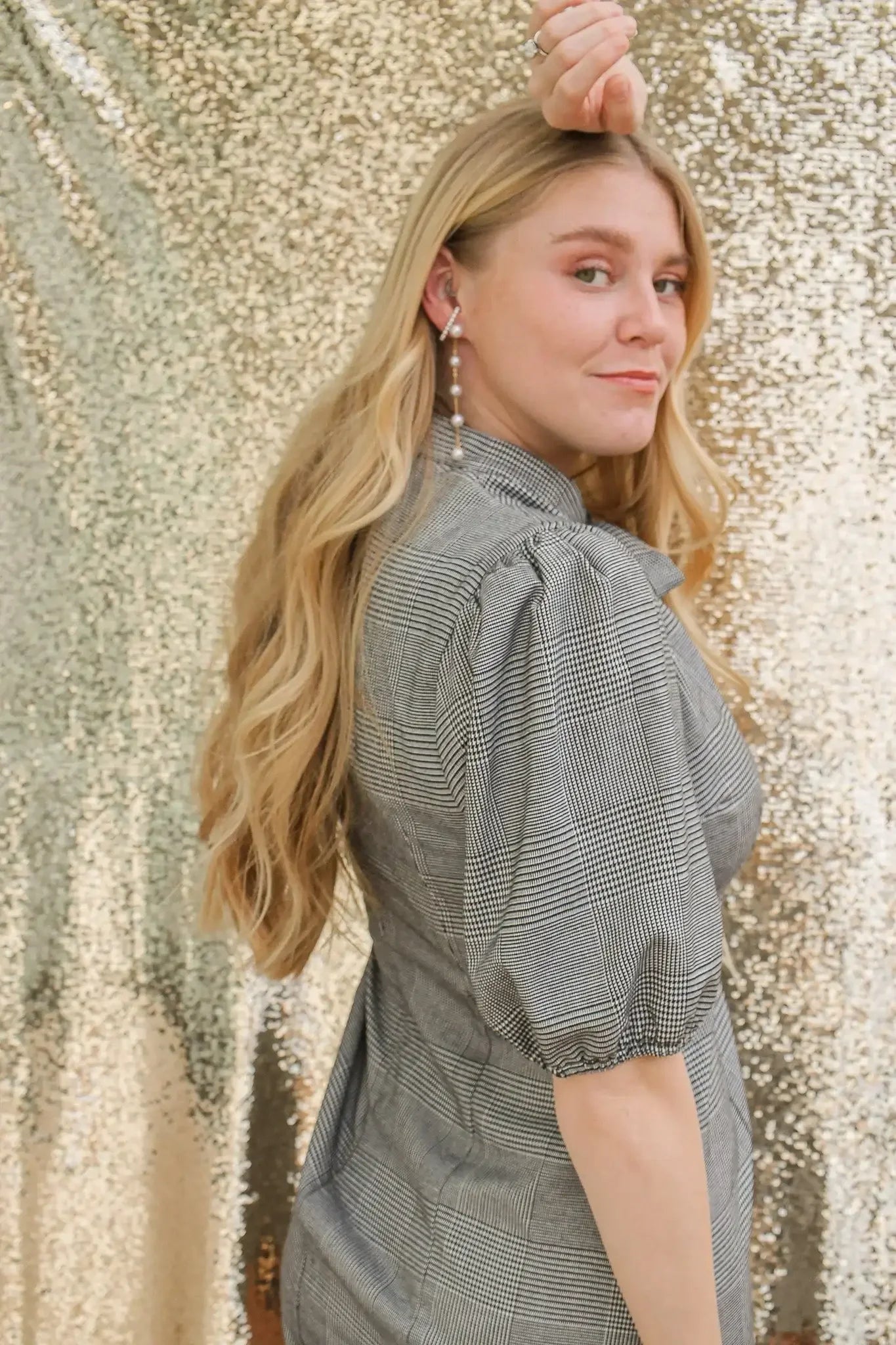 A young woman with long, wavy hair poses gracefully, her hand raised near her face. She wears a puff-sleeve, plaid dress against a shimmering gold backdrop.