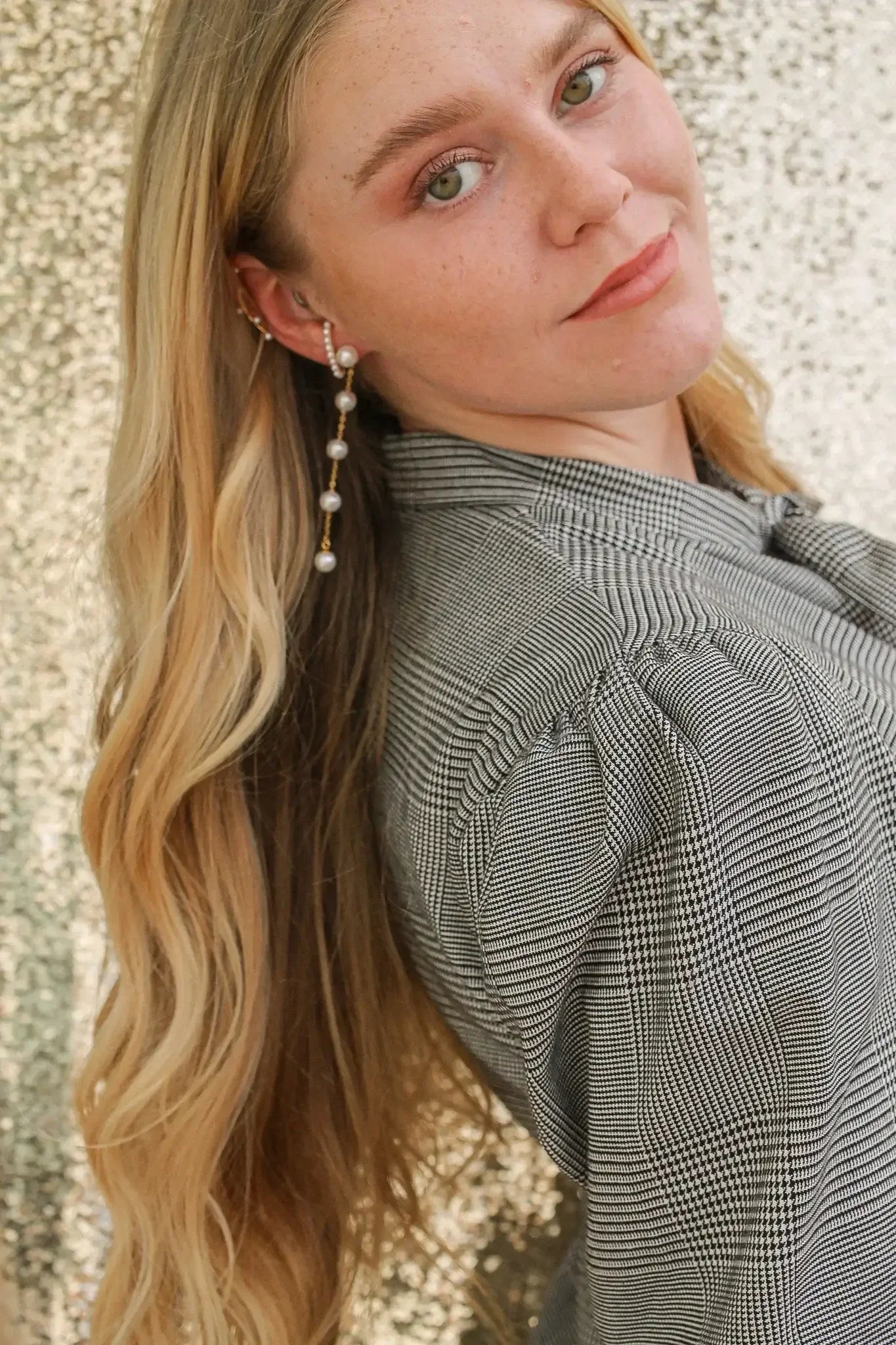 A young woman with long, wavy blonde hair is facing slightly to the side, wearing a patterned buttoned shirt. She has a pearl earring, and a sparkling backdrop enhances the scene.