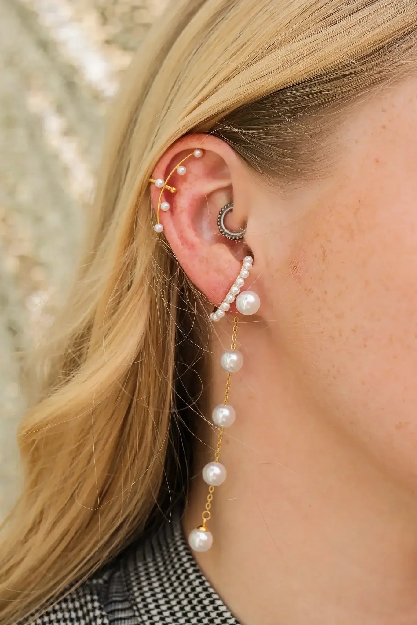 A close-up of a woman's ear adorned with multiple earrings, including a chain of pearls cascading down. The background is softly blurred, suggesting an outdoor or natural setting.