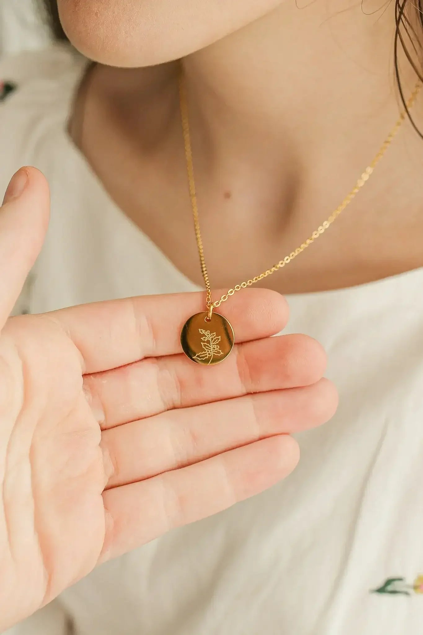 A hand holds a gold necklace pendant, depicting a floral design, while the pendant hangs near the person's neck. The background shows soft, light-colored fabric.