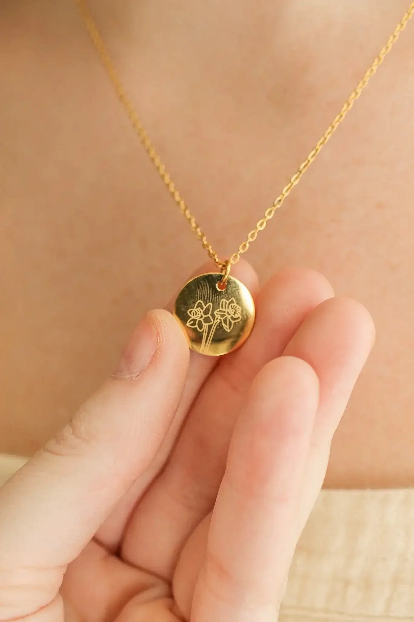 A hand holds a gold necklace featuring a round pendant engraved with flowers. The pendant hangs from a delicate chain, set against a soft, neutral background.