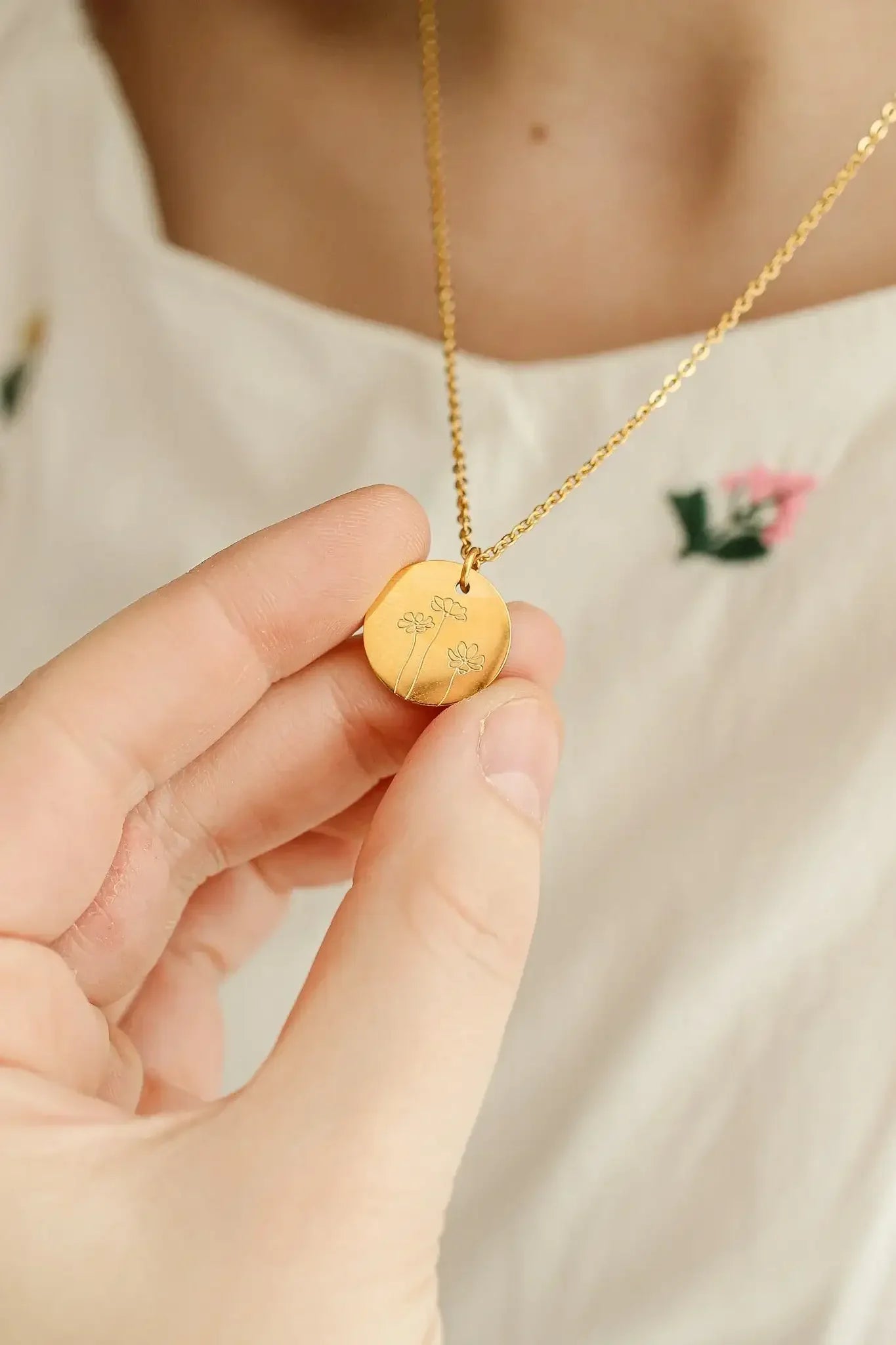 A hand holds a circular gold pendant adorned with flower engravings. The pendant hangs from a delicate chain, set against a background of a light-colored, embroidered blouse with floral accents.