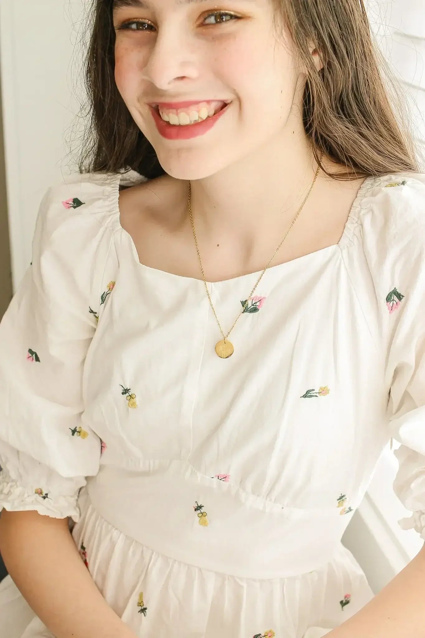 A young woman smiles, wearing a floral-embroidered white dress and a delicate necklace. She is seated near a bright window, with soft natural light illuminating her face.