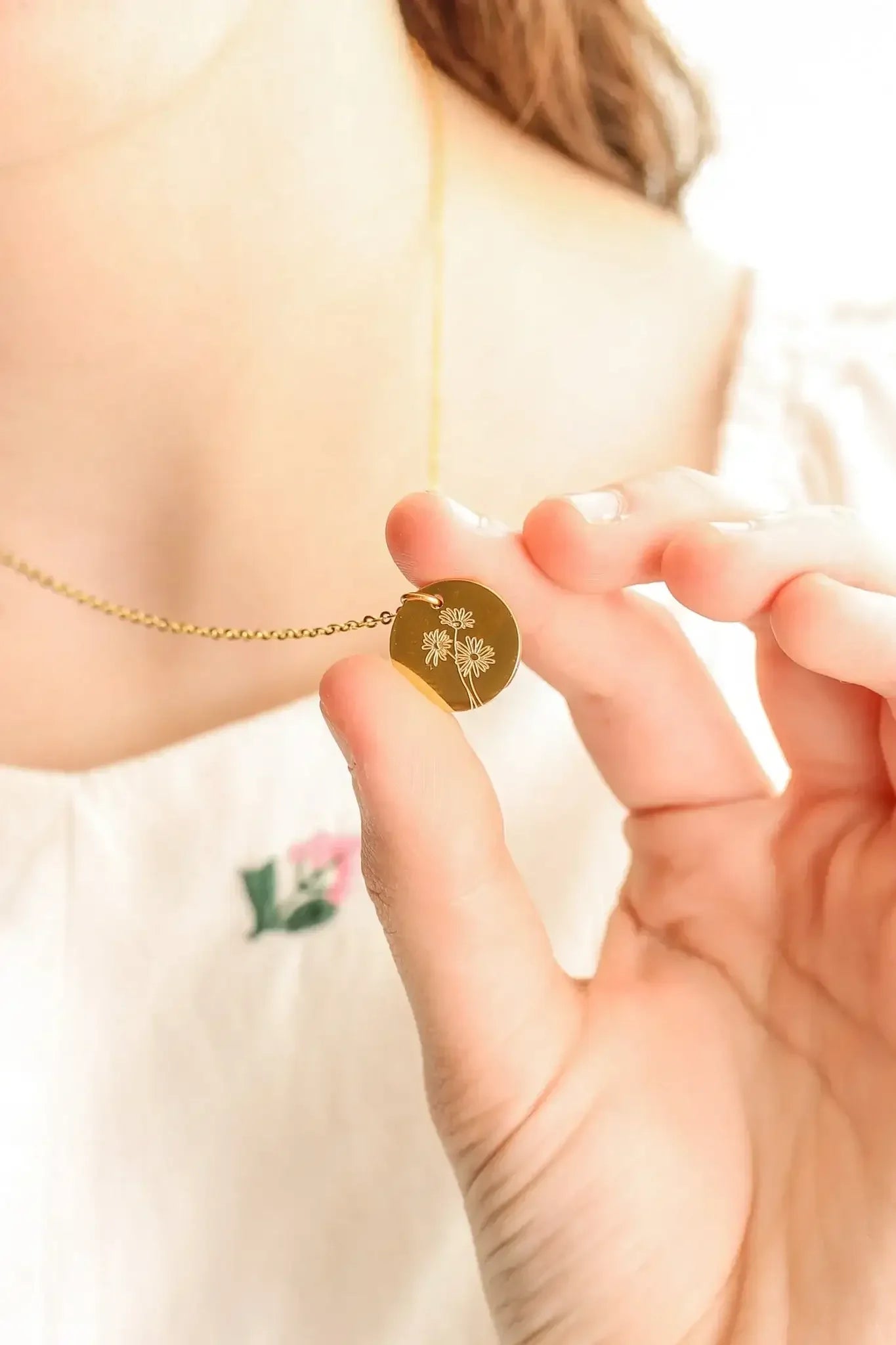 A hand holds a circular gold necklace pendant with an engraved floral design, while a person’s collarbone and part of a light-colored garment are visible in a soft, bright environment.