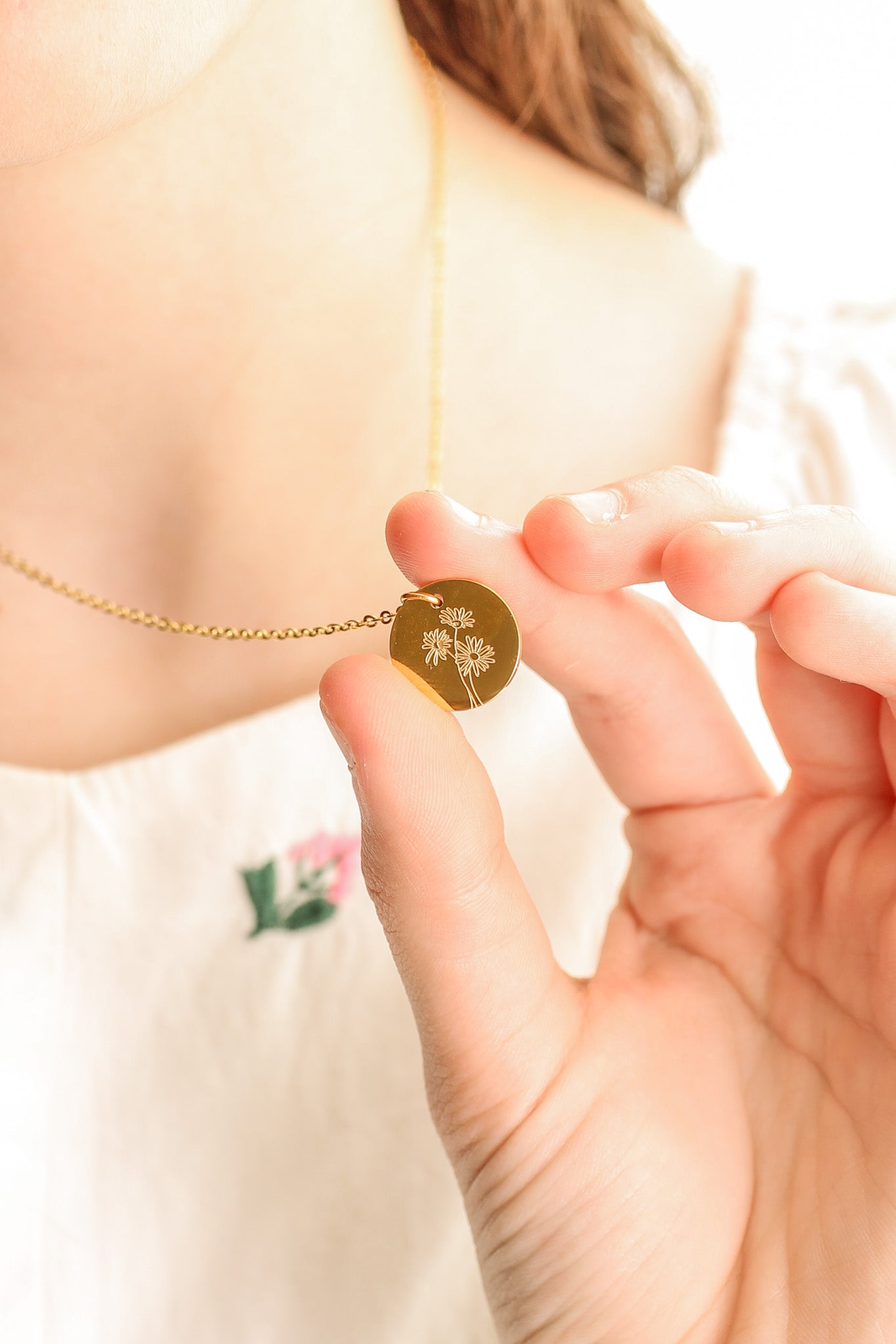 A hand holds a circular gold necklace pendant with an engraved floral design, while a person’s collarbone and part of a light-colored garment are visible in a soft, bright environment.