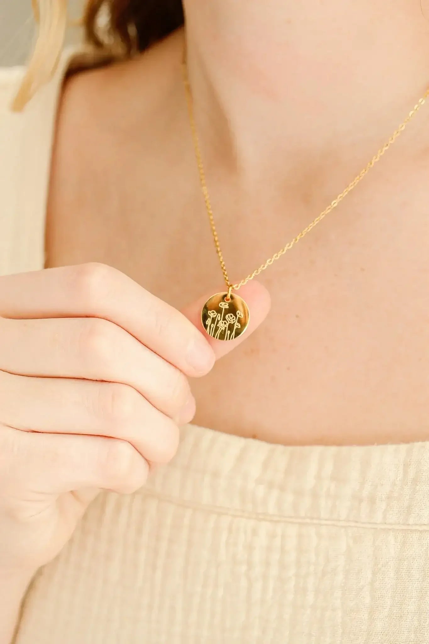 A person holds a gold pendant featuring floral engravings, showcasing it against a textured, light-colored fabric background, highlighting the pendant's design and the gentle softness of the attire.