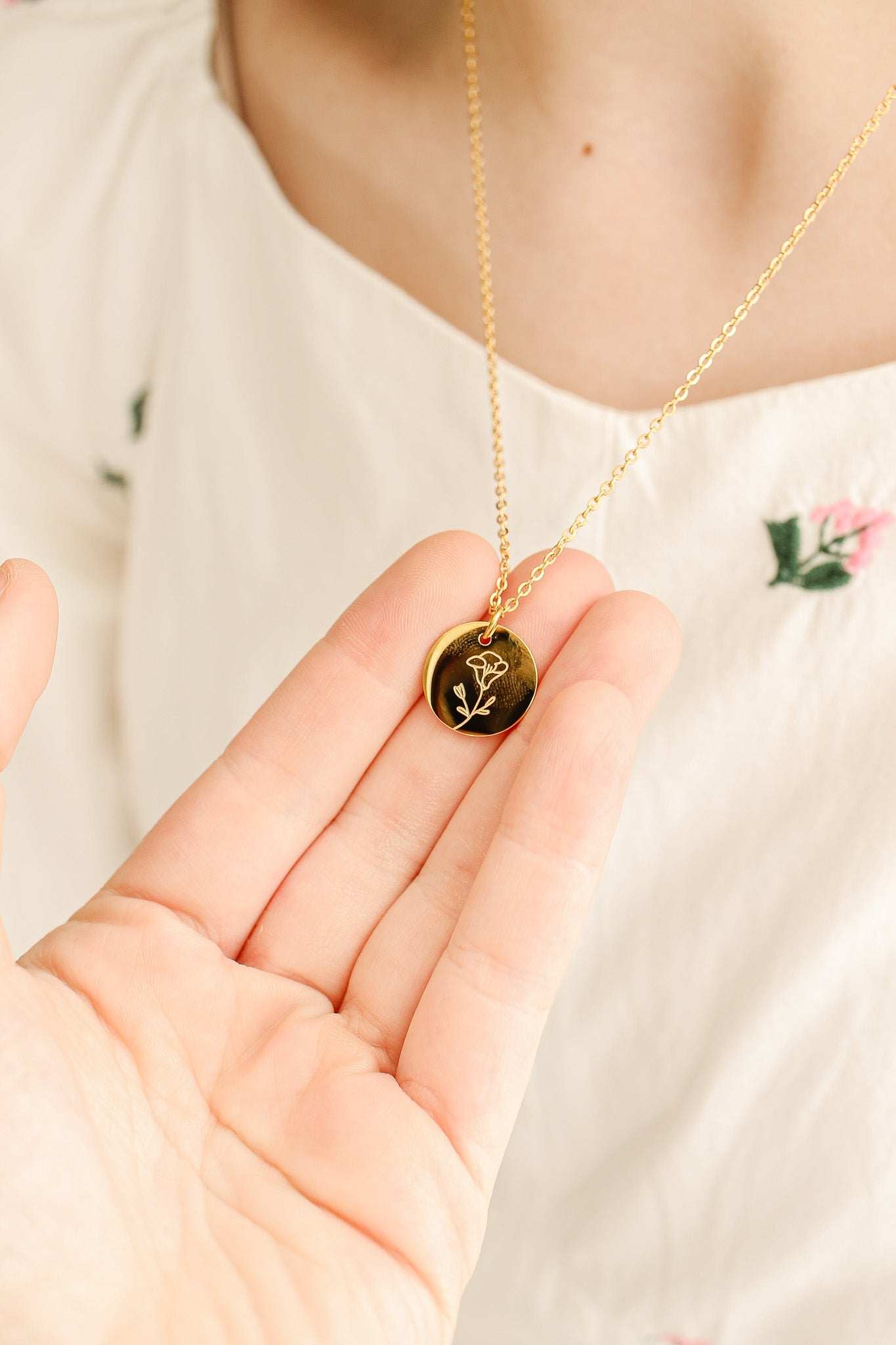 A hand holds a gold pendant featuring an engraved flower design. The person wearing a light, floral-embroidered dress is showcasing the necklace against a softly lit, neutral background.