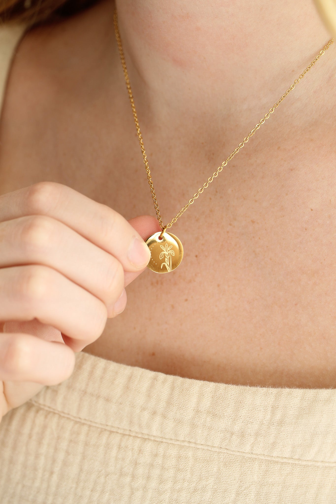 A person holds a gold pendant necklace featuring a floral design, displaying it against a softly lit, neutral-toned background that reveals part of their skin and clothing.