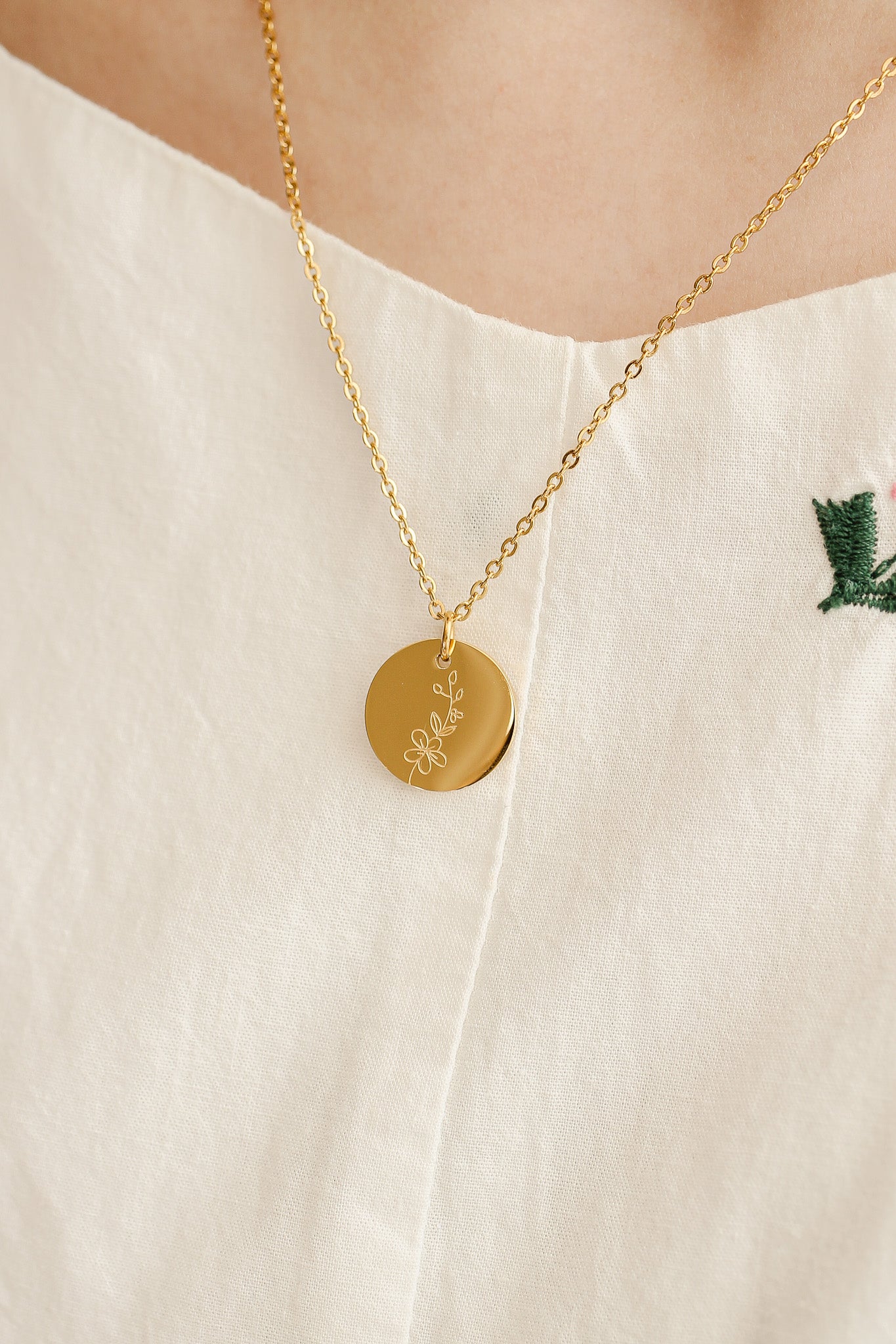 A gold necklace with a circular pendant featuring a floral design hangs on a person's neck, showcasing delicate chain links against a light-colored, textured garment.