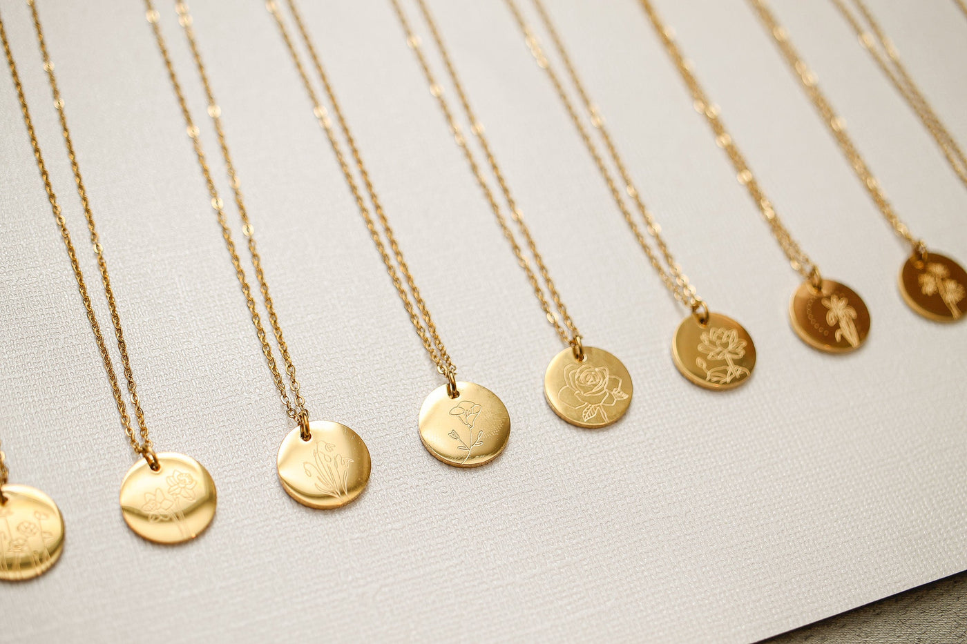 Gold necklaces hang in a row with engraved circular pendants, showcasing various floral designs. They are displayed on a textured light-colored background, emphasizing their elegance.
