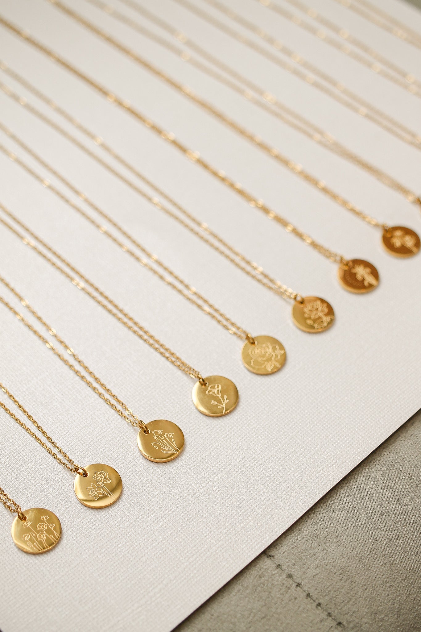 Gold necklaces hang in a straight line on a textured surface. Each necklace features a circular pendant engraved with floral designs, showcasing an elegant display of jewelry.