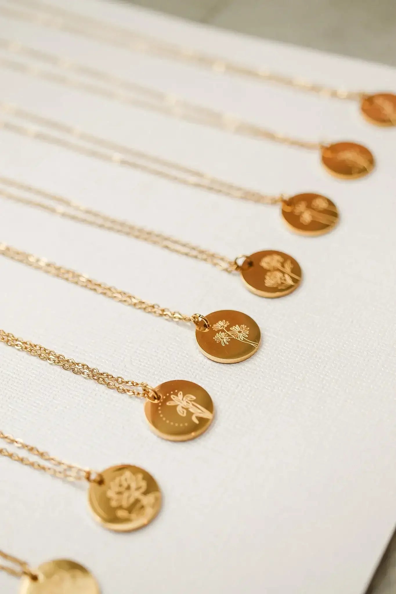Gold necklaces with circular pendants are displayed side by side. Each pendant features unique floral engravings, resting on a textured light-colored background.