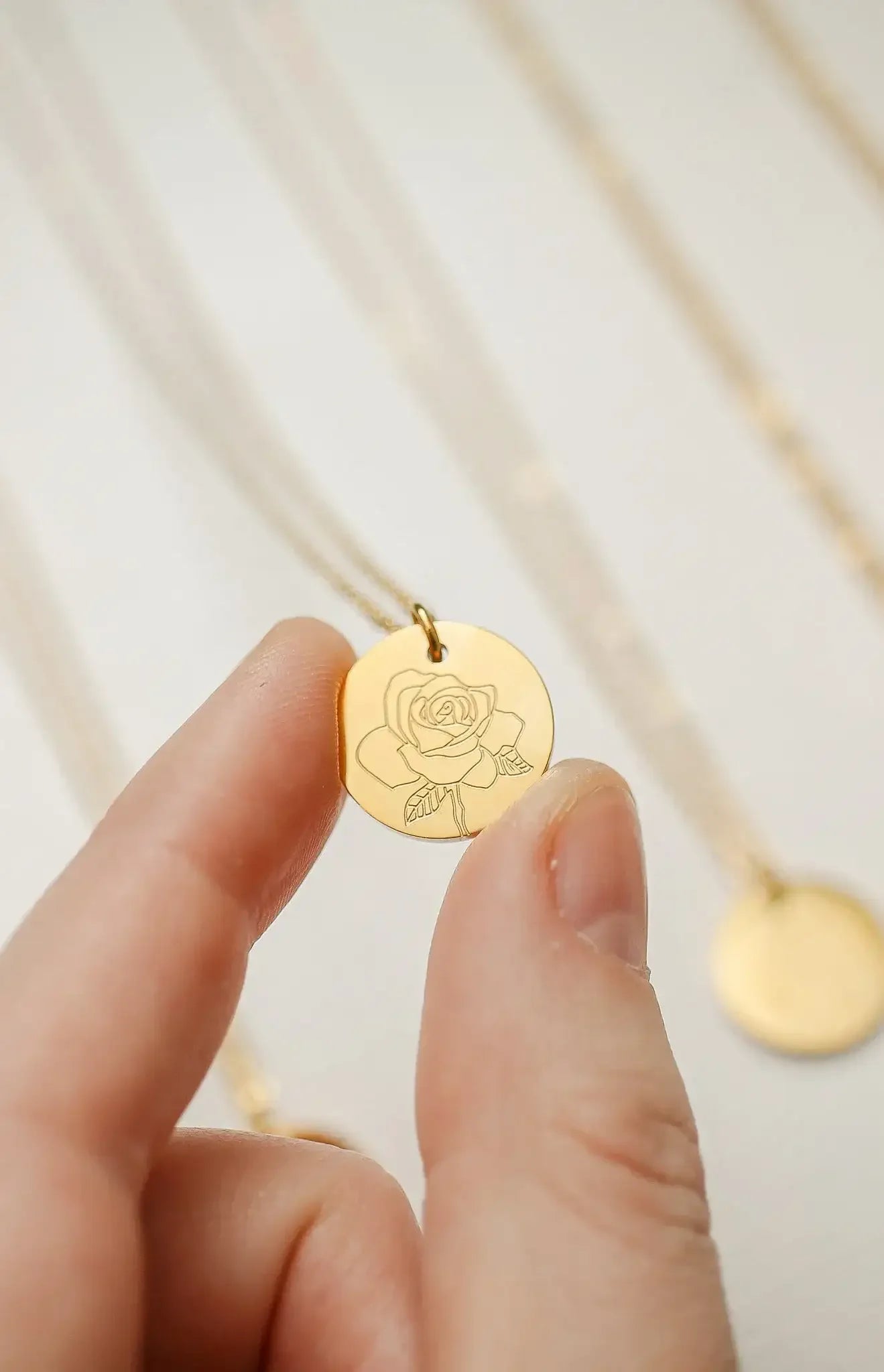 A hand holds a gold pendant featuring an engraved rose design. Multiple delicate gold chains are visible in the background against a light, neutral surface.