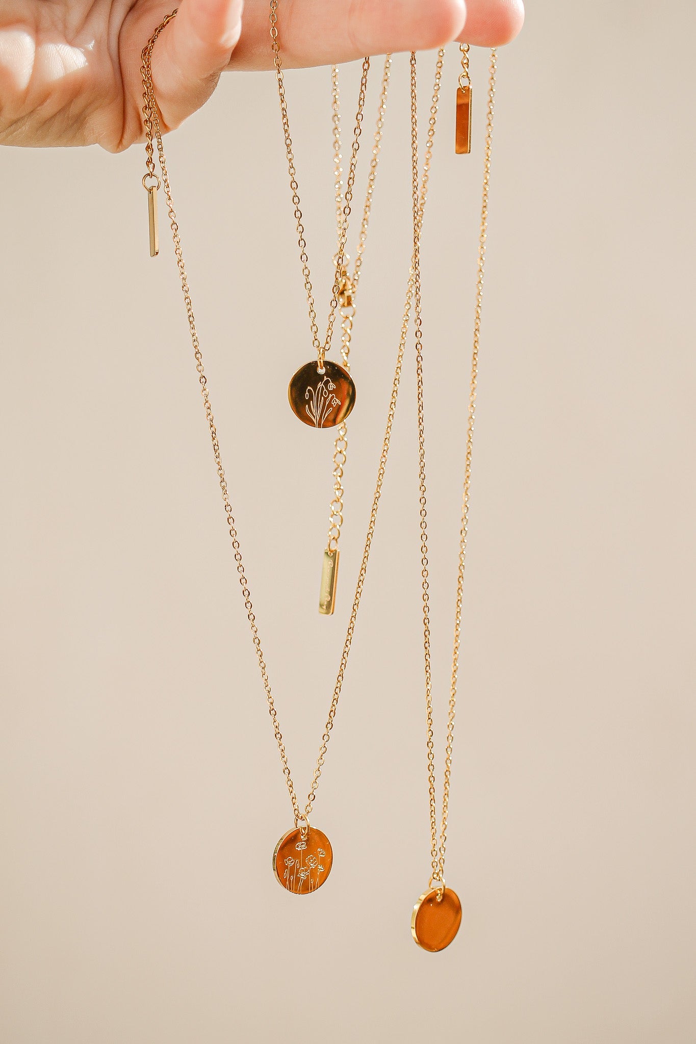 A hand holds several gold necklaces, showcasing various pendants including circular ones with floral designs and small rectangular charms, against a soft, neutral background.
