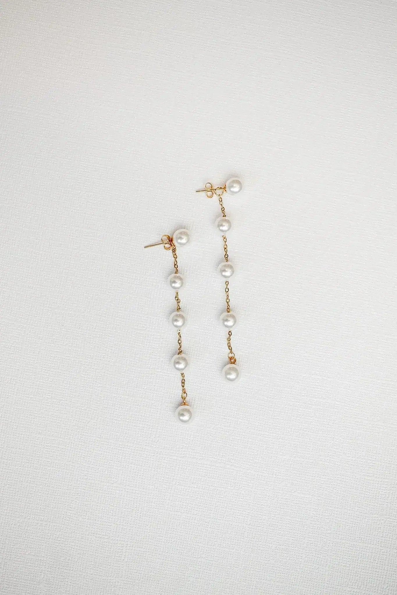 Two elegant gold earrings are displayed, featuring multiple dangling pearls connected by delicate chains. They rest on a textured white background, emphasizing their sophistication and shine.