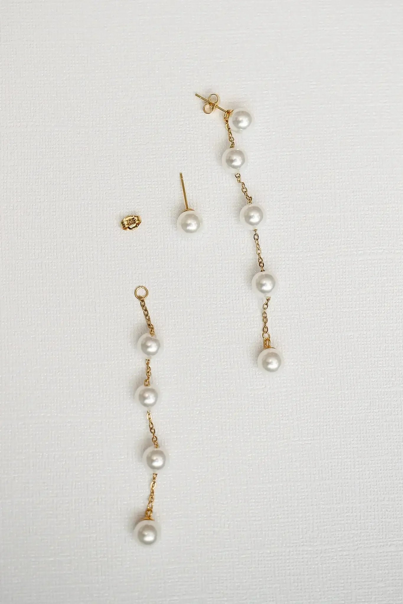 Pearl earrings, featuring a gold chain with multiple pearls, rest on a textured white background. A small gold backing sits nearby, emphasizing the elegance of the jewelry arrangement.