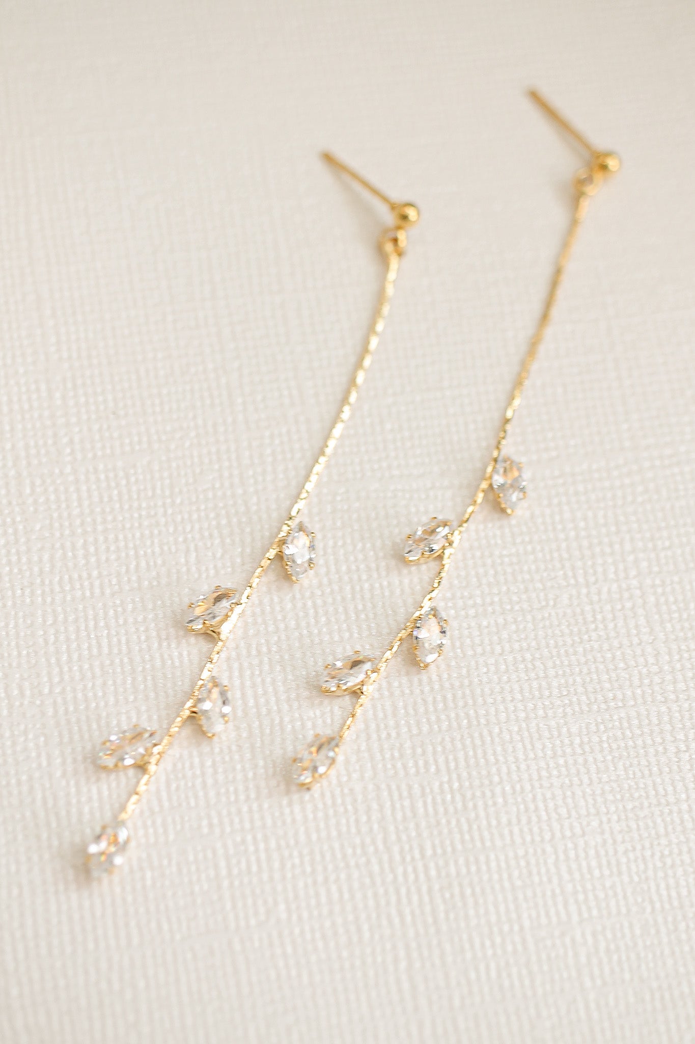 Two gold earrings with delicate, vine-like designs hang gracefully. They feature small, sparkling crystals arranged in a linear pattern. The background is a simple, textured cream surface.