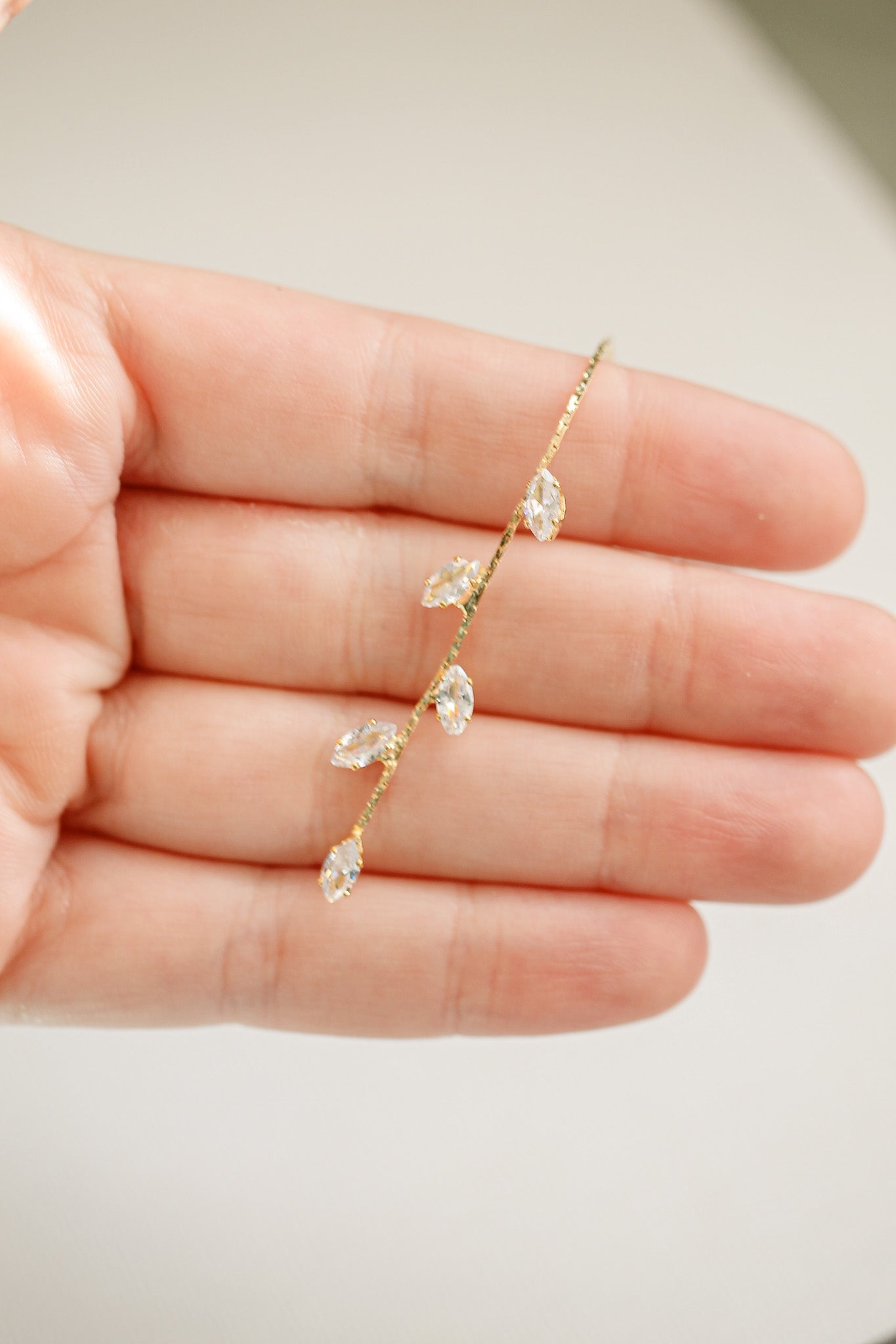 A hand holds a delicate hairpin shaped like a branch, adorned with sparkling leaf-like crystals. The background is softly blurred, emphasizing the intricate design of the accessory.