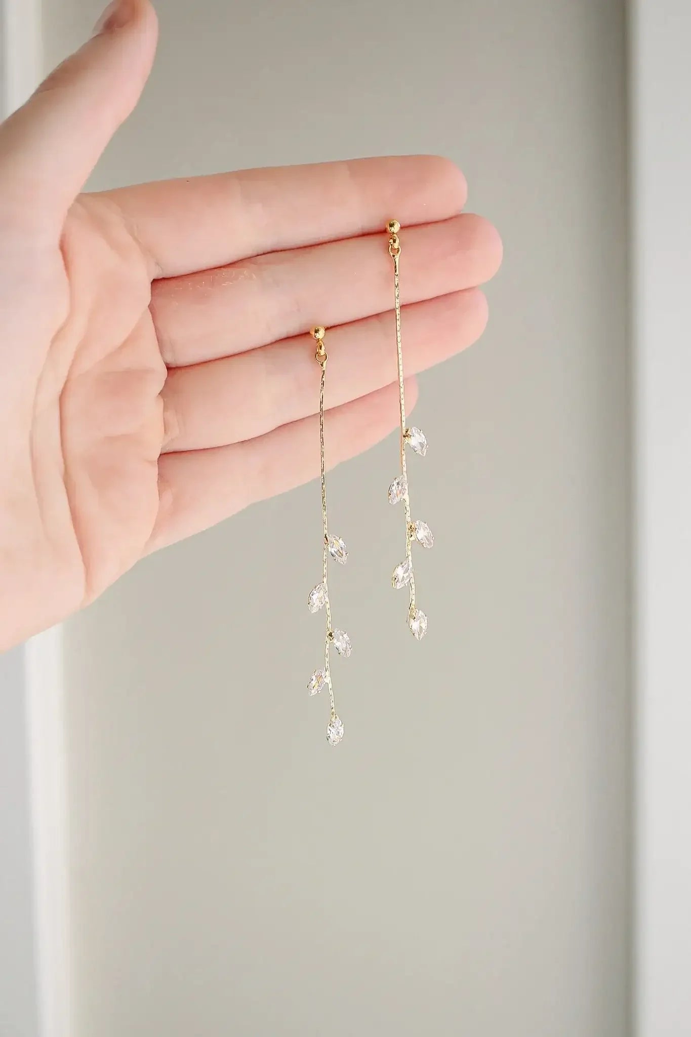 A hand holds a pair of elegant, dangling gold earrings adorned with tiny, sparkling crystals shaped like leaves, against a soft, neutral background.