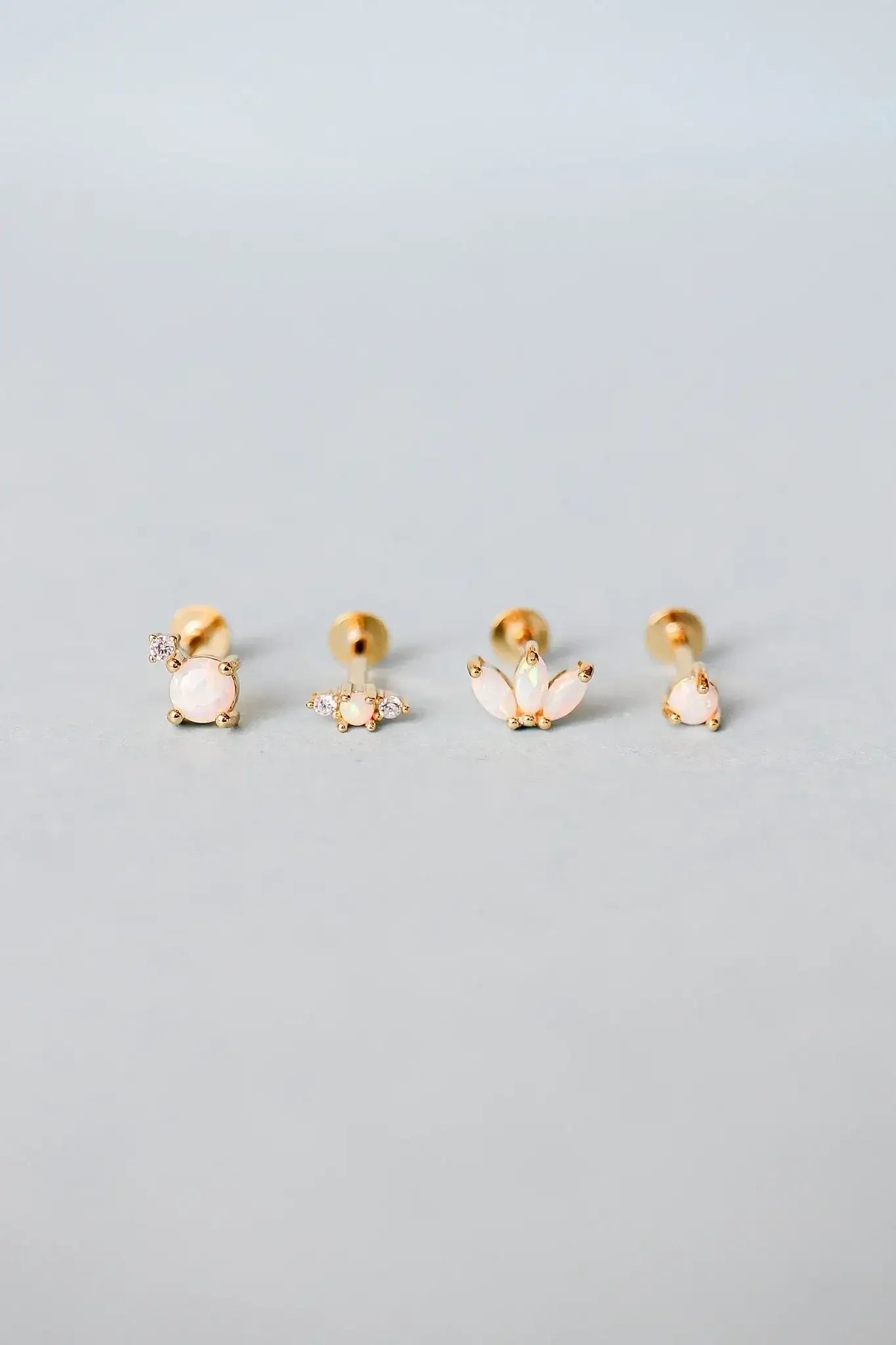 Four pairs of gold stud earrings, each featuring different designs with opal-like stones, are arranged neatly on a soft gray background. The earrings display a mix of shapes and embellishments.