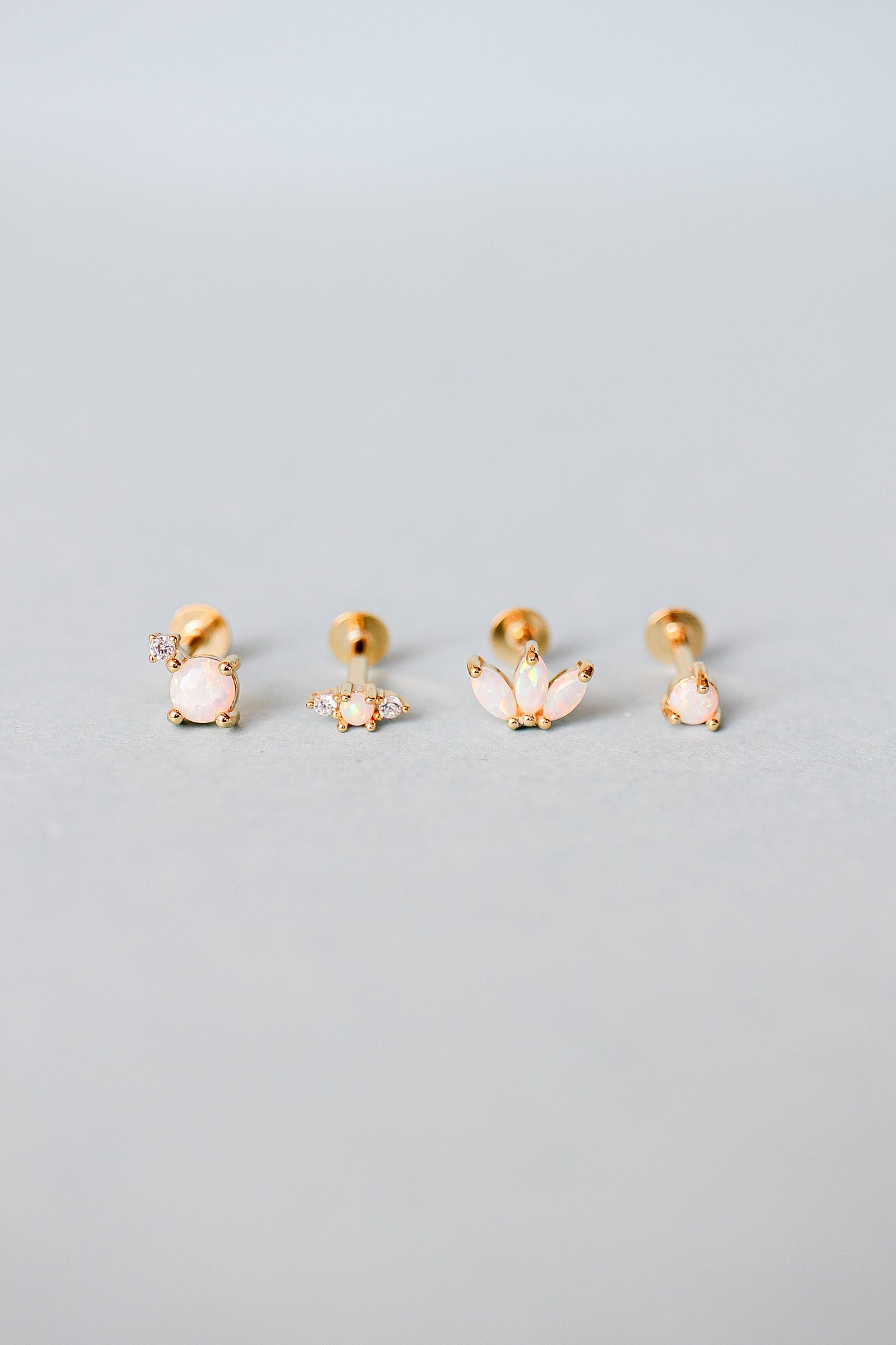 Four pairs of gold stud earrings, each featuring different designs with opal-like stones, are arranged neatly on a soft gray background. The earrings display a mix of shapes and embellishments.