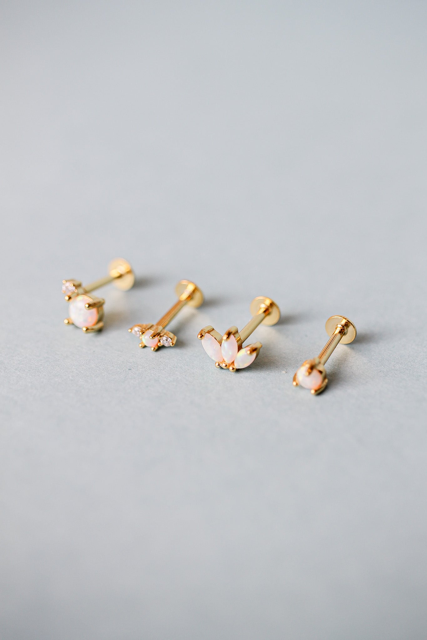 Gold earrings with opal-like stones are arranged in a row on a smooth, light gray surface. Their elegant designs vary, showcasing floral and geometric patterns.