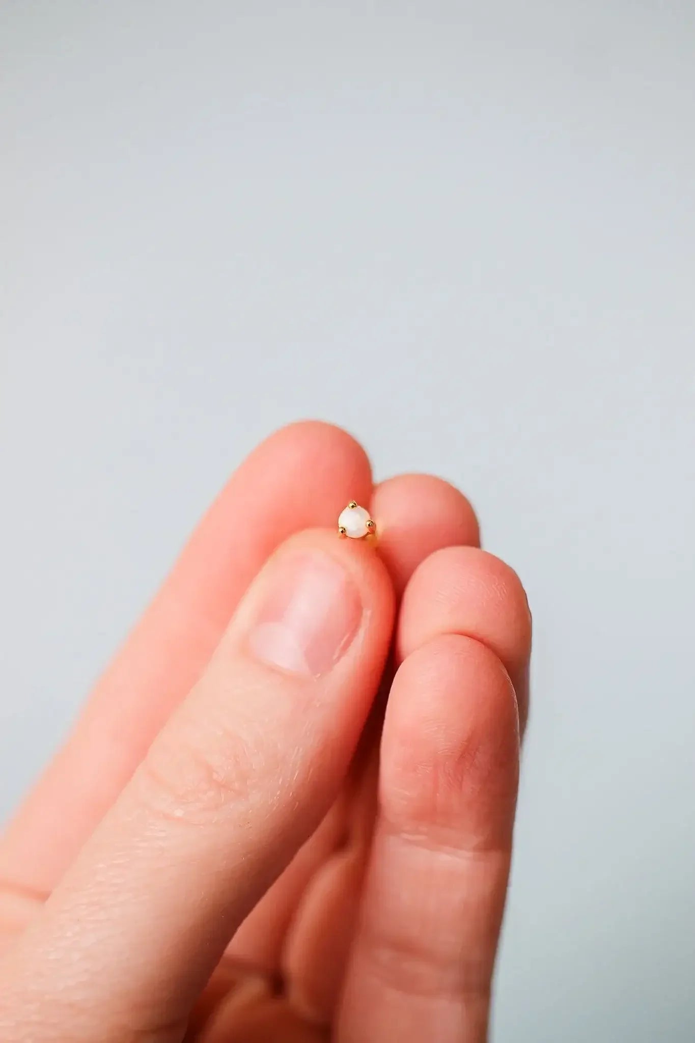 A hand holds a small, delicate earring featuring a translucent stone set in gold. The background is a soft, muted color, enhancing the focus on the jewelry.
