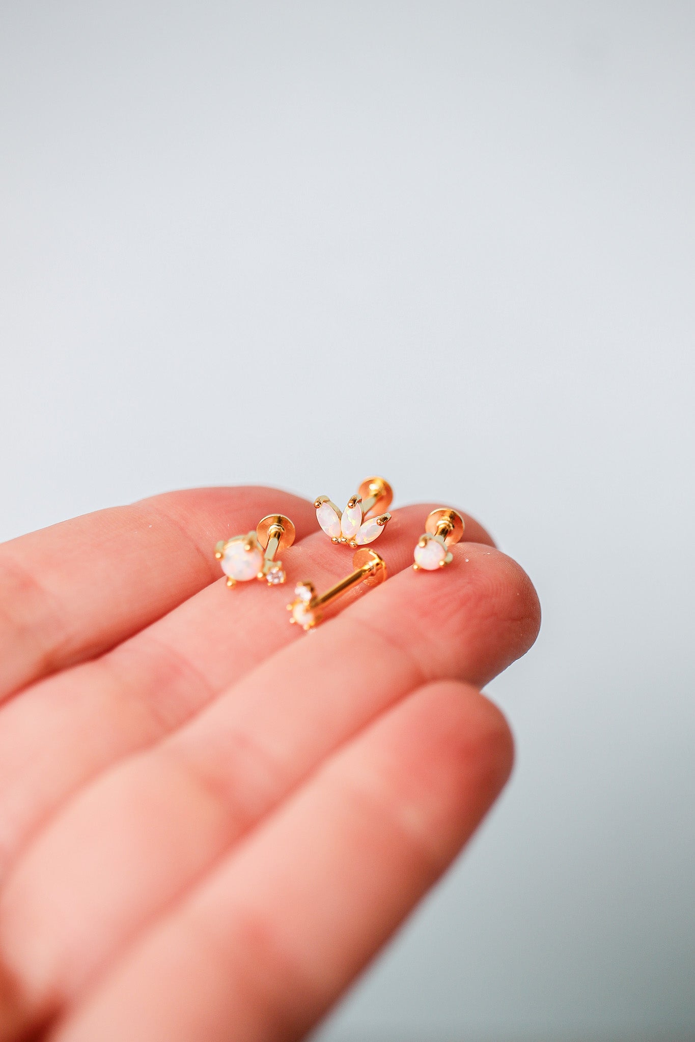A hand holds several gold earrings, featuring opal and rhinestone details, against a soft, light background. The jewelry is delicately arranged, showcasing its design and shimmer.