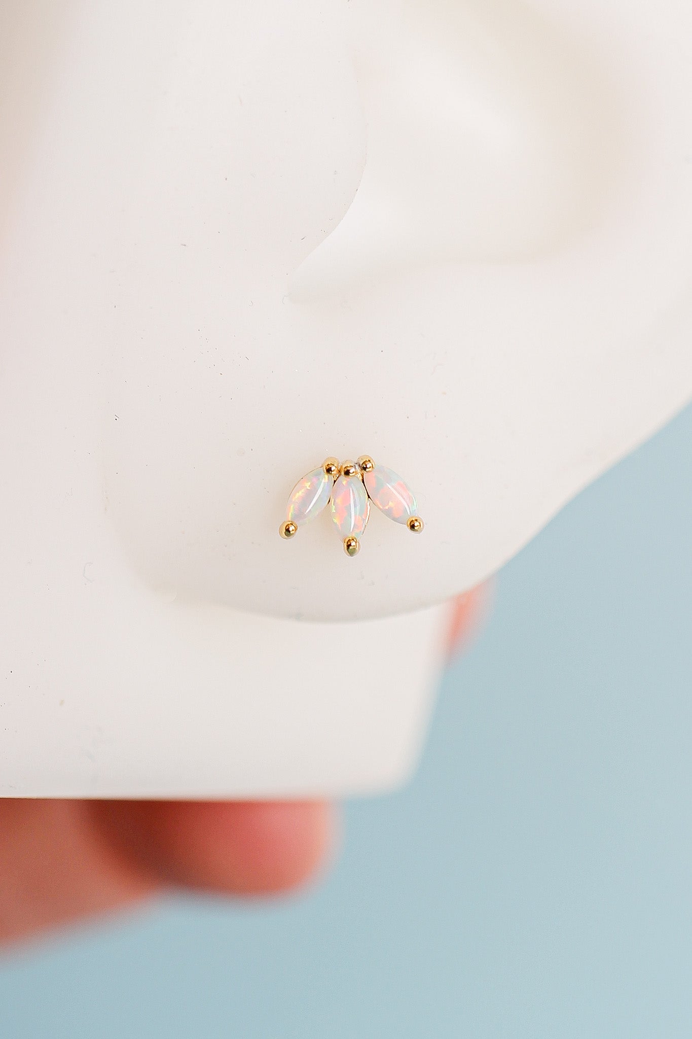 A small gold earring featuring three opalescent, leaf-shaped stones is displayed on a mannequin ear, with a blurred hand in the background, suggesting a setting focused on jewelry presentation.