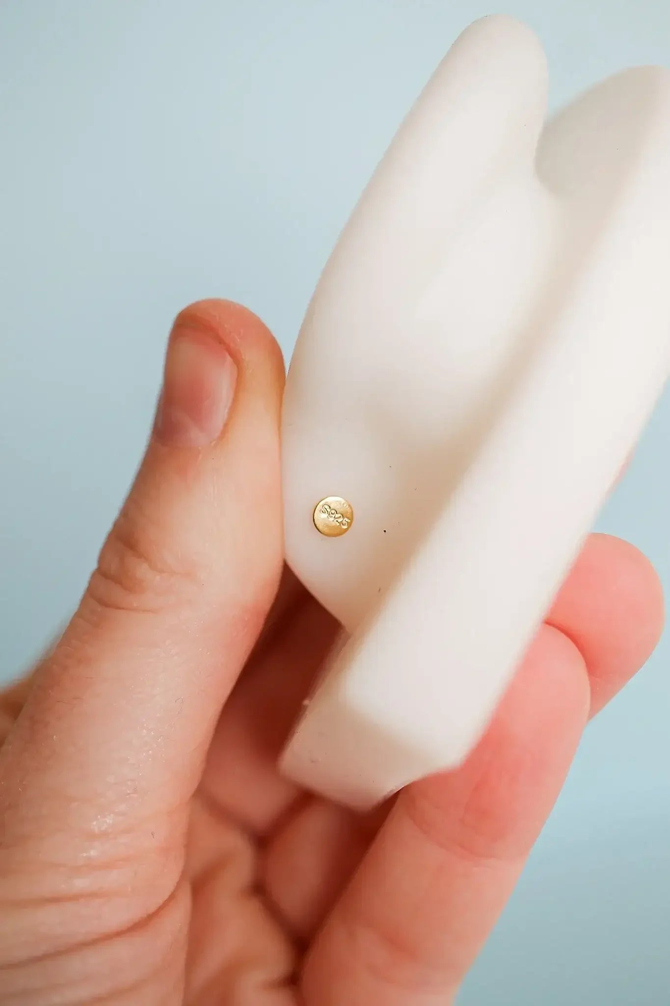 A hand holds a white silicone object, which features a small gold circular decoration. The background is a soft blue, creating a calm and minimalist setting.