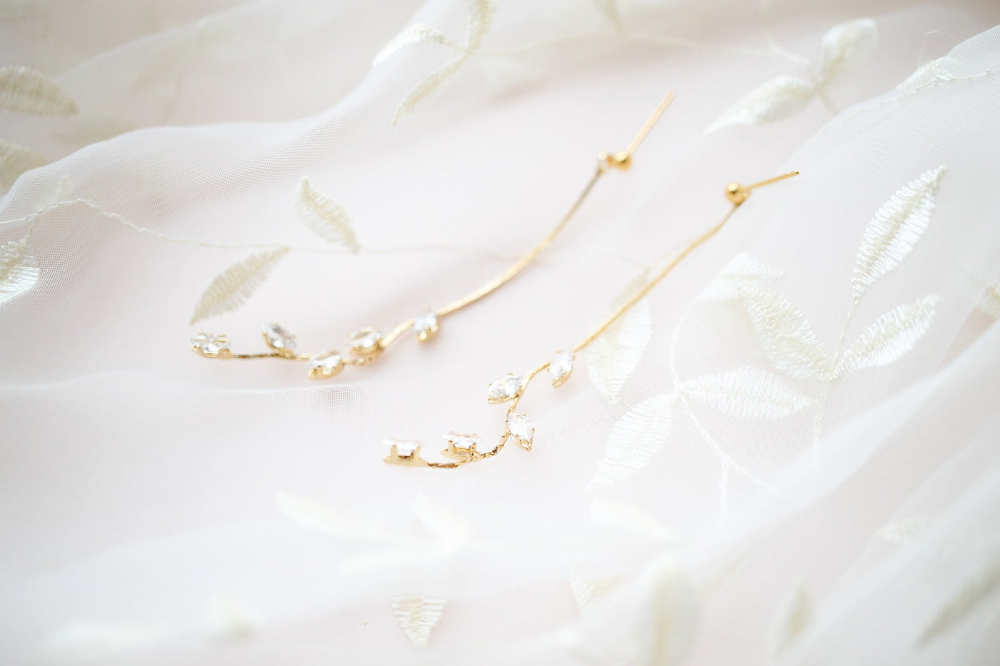 Delicate gold earrings, designed in a floral vine pattern with sparkling gemstones, rest atop soft, sheer fabric embroidered with leaf motifs, creating an elegant and romantic display.