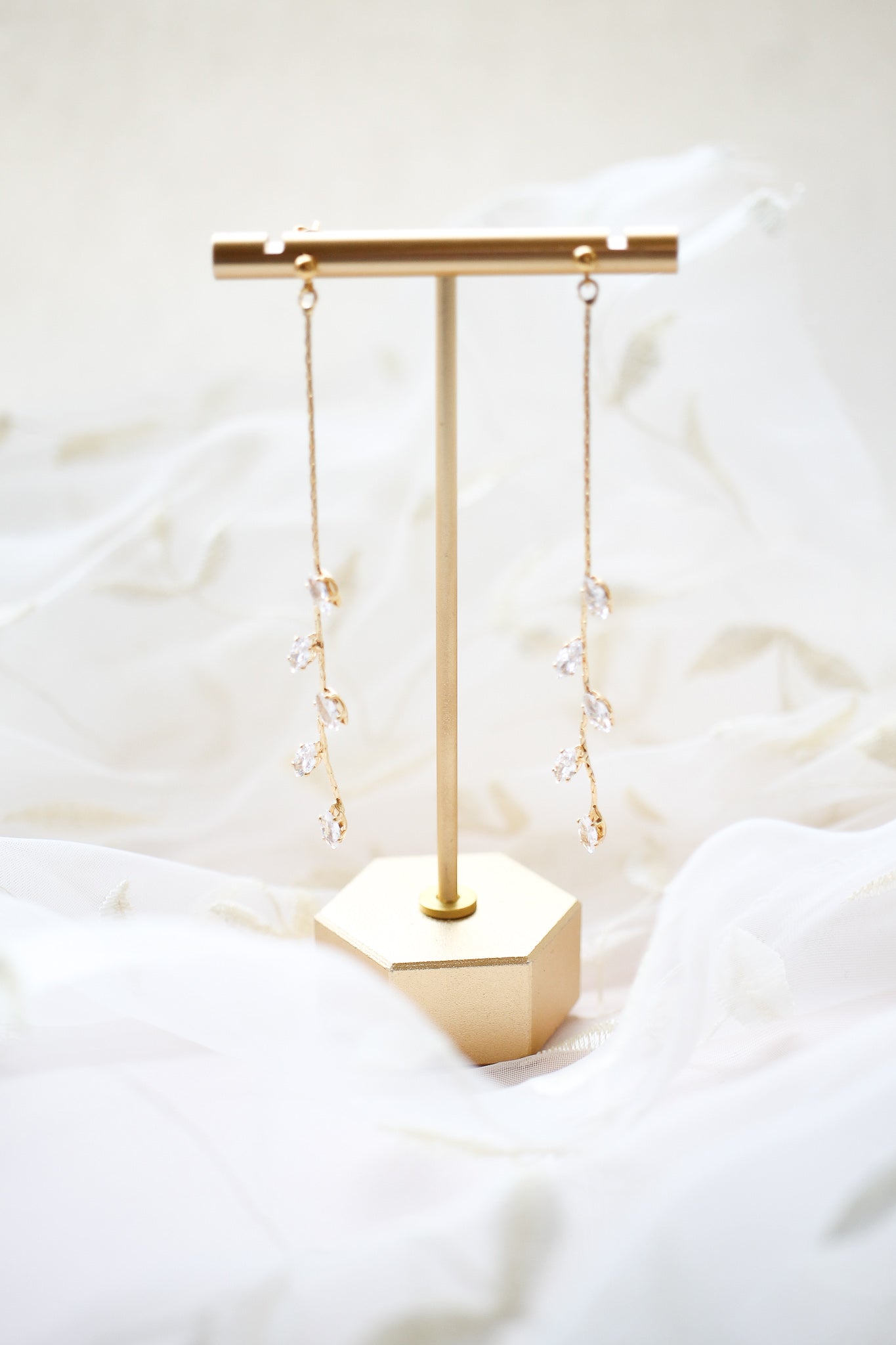 A pair of elegant, dangling earrings with crystal-like embellishments hang from a minimalist gold display stand, set against a soft, flowing white fabric background.