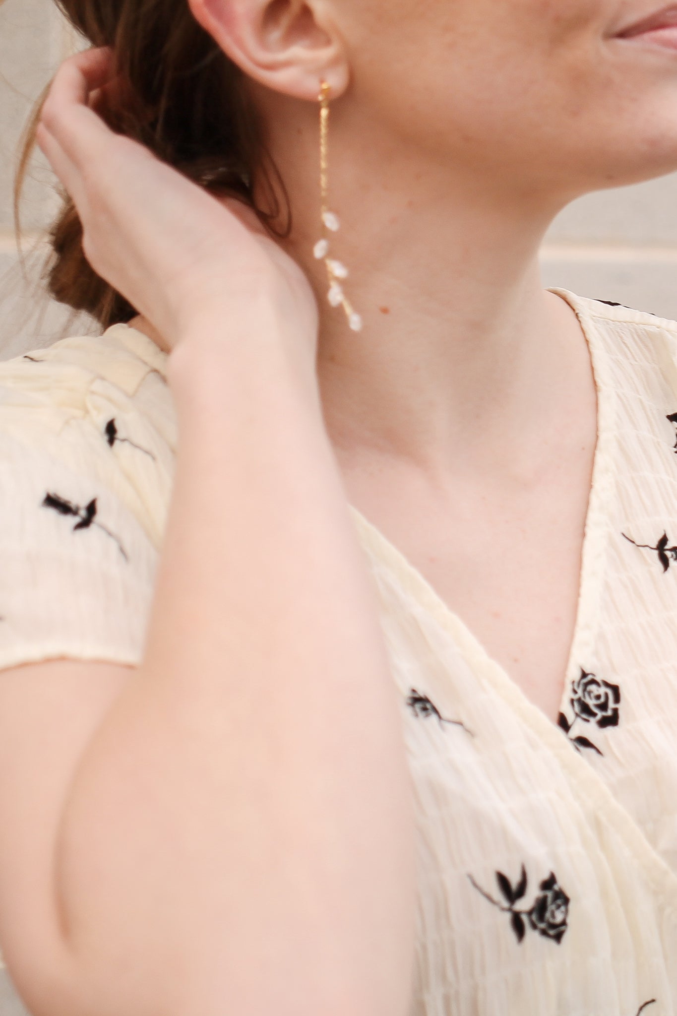 A woman wears a detailed, long gold earring with floral accents, gently touching her hair. She is dressed in a light, patterned blouse against a softly blurred neutral background.