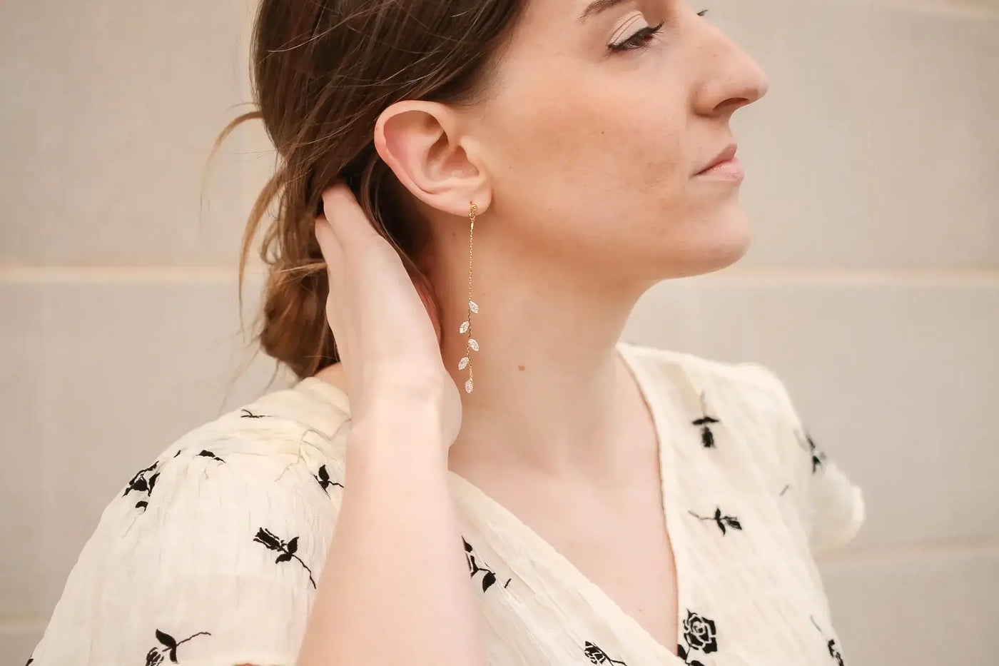A woman with long hair, wearing a light floral blouse, gently touches her neck while wearing a dangling leaf-patterned earring, set against a neutral background.