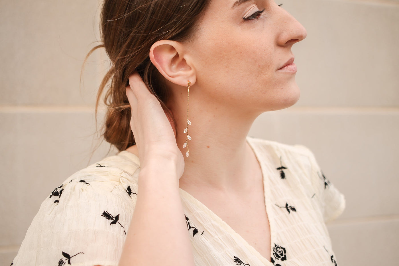 A woman with long hair, wearing a light floral blouse, gently touches her neck while wearing a dangling leaf-patterned earring, set against a neutral background.