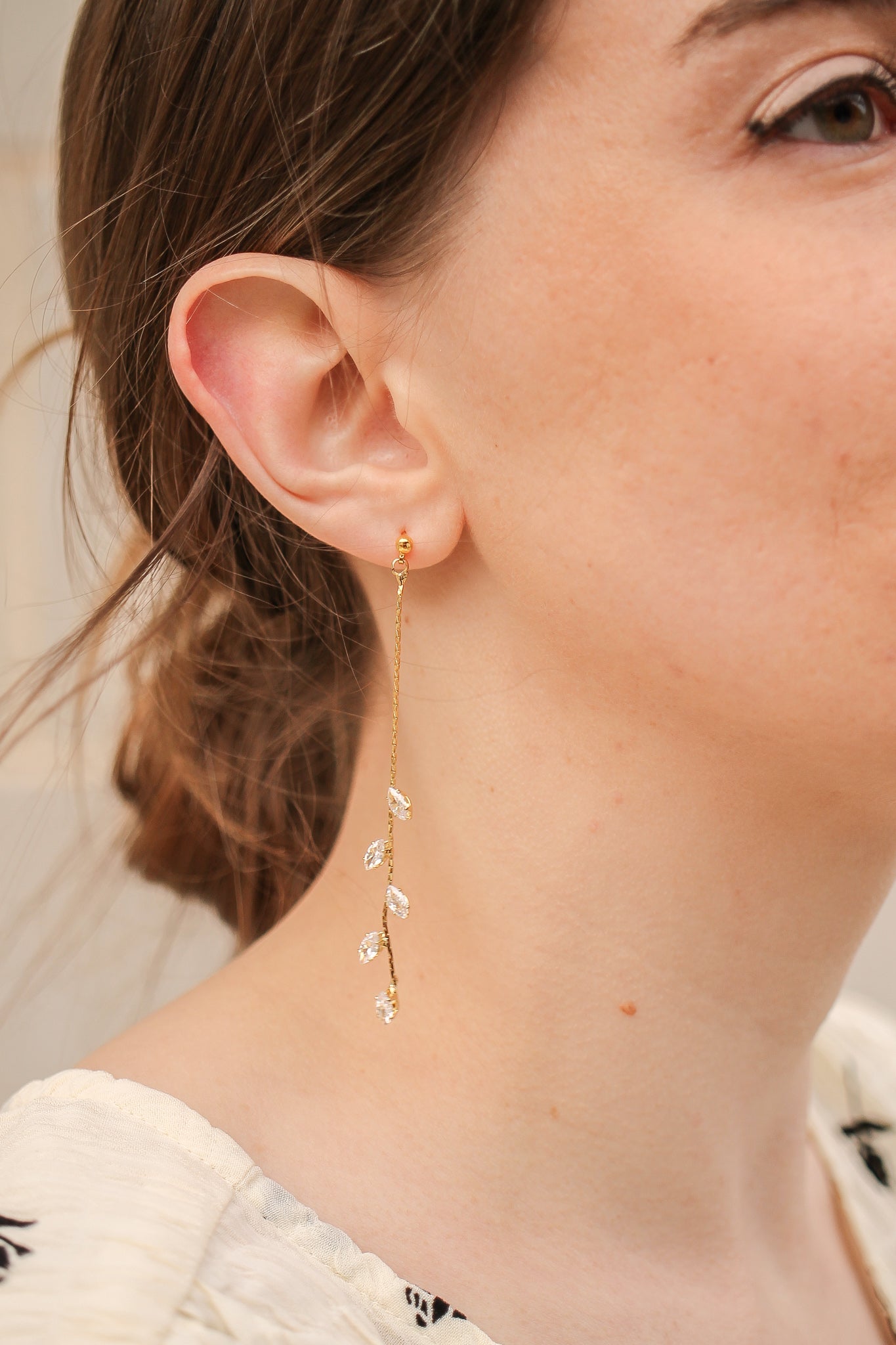 A gold dangling earring with small, leaf-shaped decorations is showcased against the profile of a woman. She has light brown hair styled loosely, and a soft, natural background.