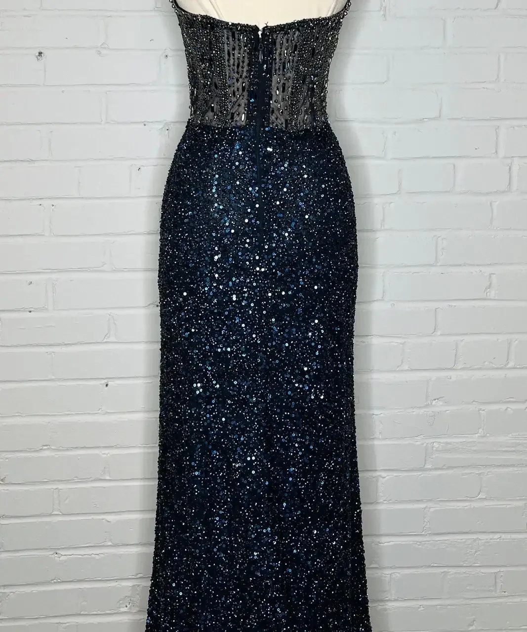 A shimmering, floor-length navy gown adorned with sequins and a transparent bodice is displayed on a dress form against a white brick wall background. The dress exudes elegance and glamour.