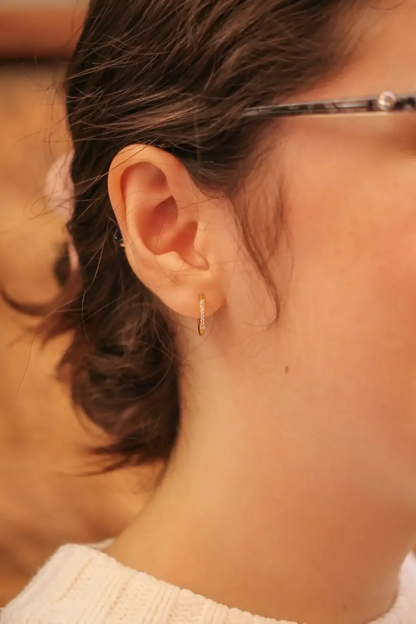 A close-up of a woman's ear features a small gold hoop earring, with her hair styled in loose waves. She is wearing a light-colored knitted sweater, set against a softly blurred background.