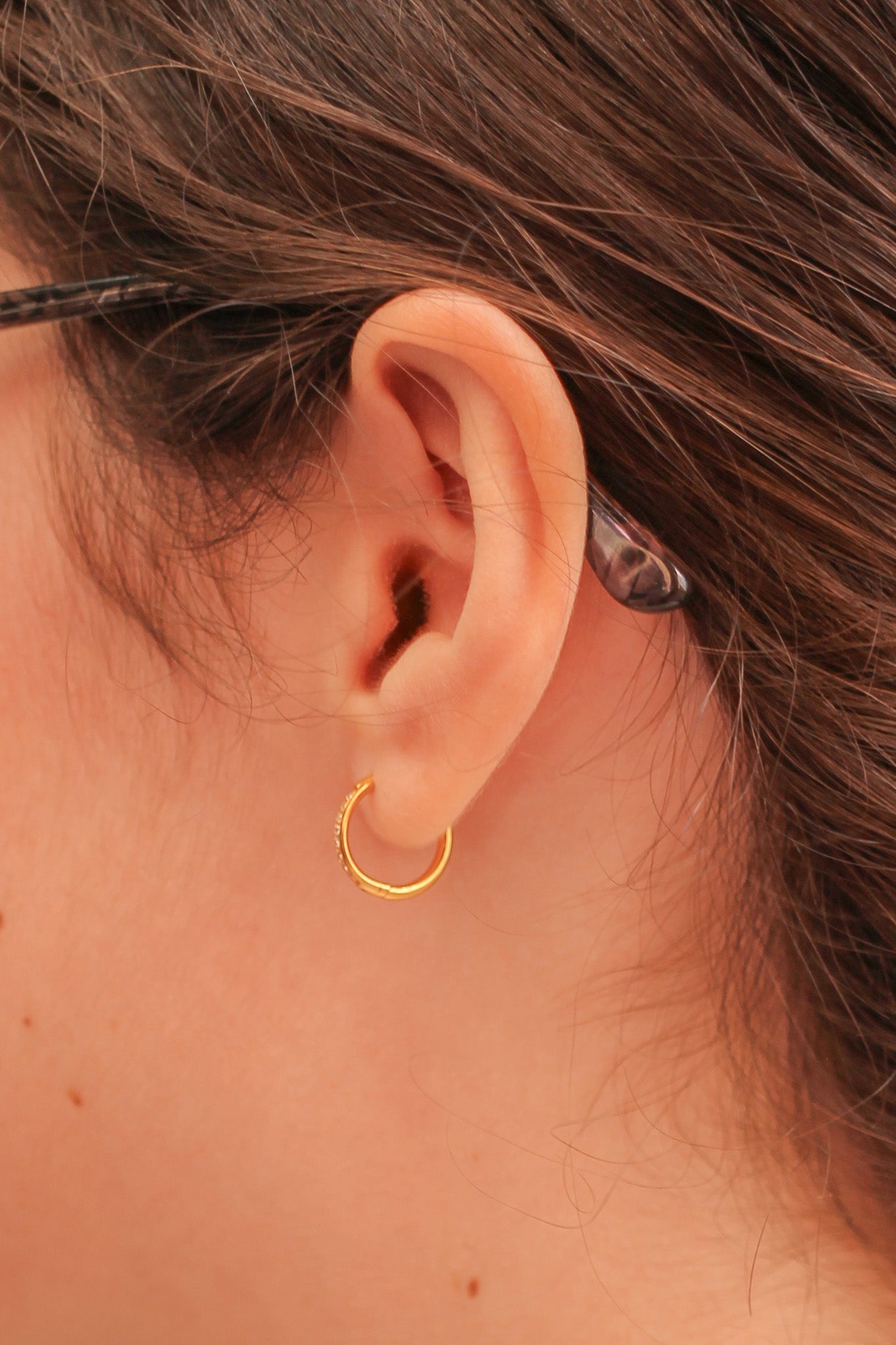 A close-up of a human ear adorned with a small gold hoop earring, surrounded by brown hair, creating an intimate view of jewelry in a personal context.